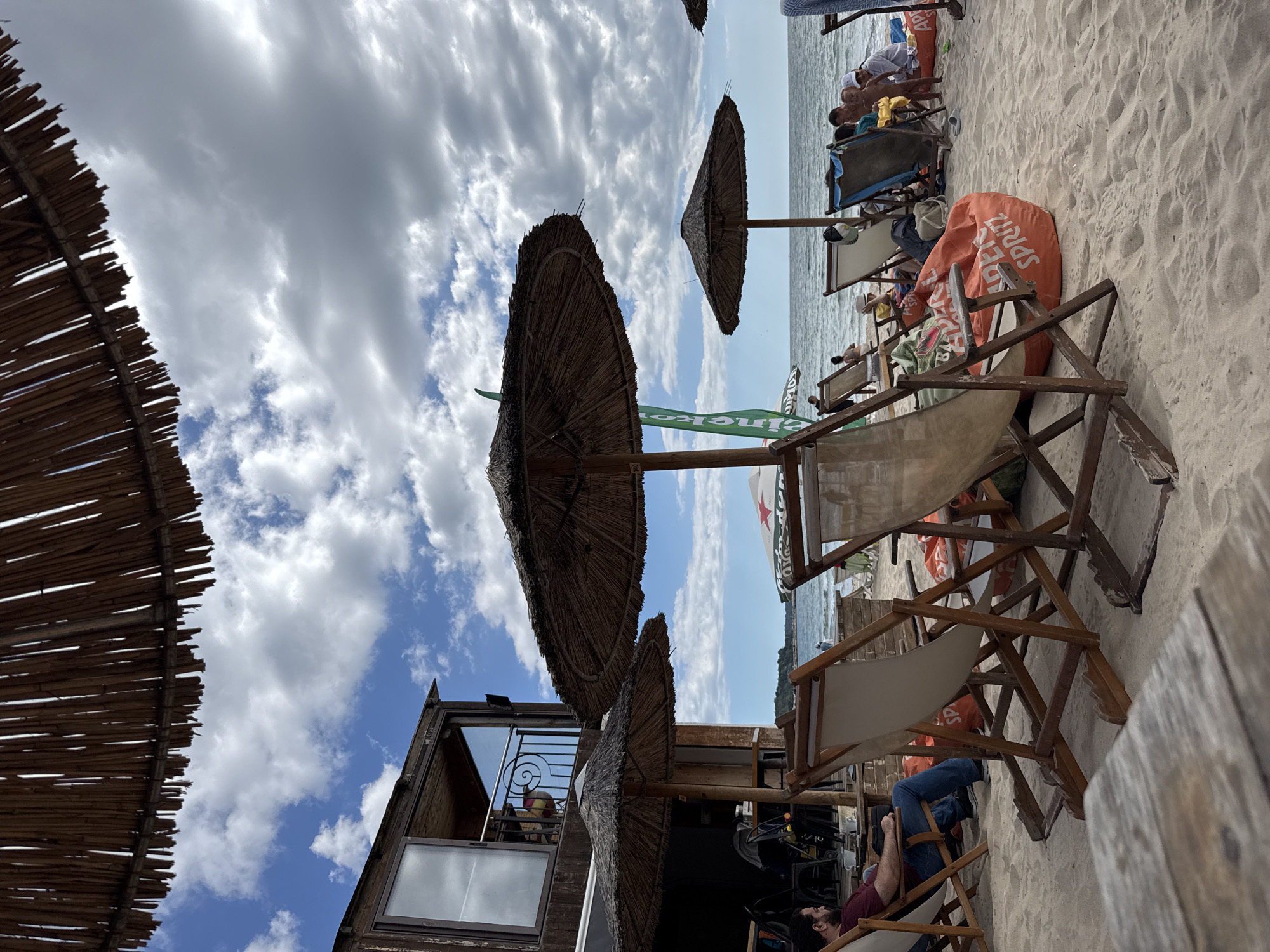 The image depicts a beach scene at a cafe or bar in Varna, Bulgaria. The foreground features a wooden table, and beyond it, several beach chairs are set up on the sand. One man is seated in a chair, looking towards the sea. In the background, people are relaxing on the beach and in chairs, with the sea and a distant shoreline visible. Several straw umbrellas provide shade. A two-story wooden building, likely part of the establishment, is visible on the left side of the frame. The sky is partly cloudy, with a mix of blue sky and white clouds.