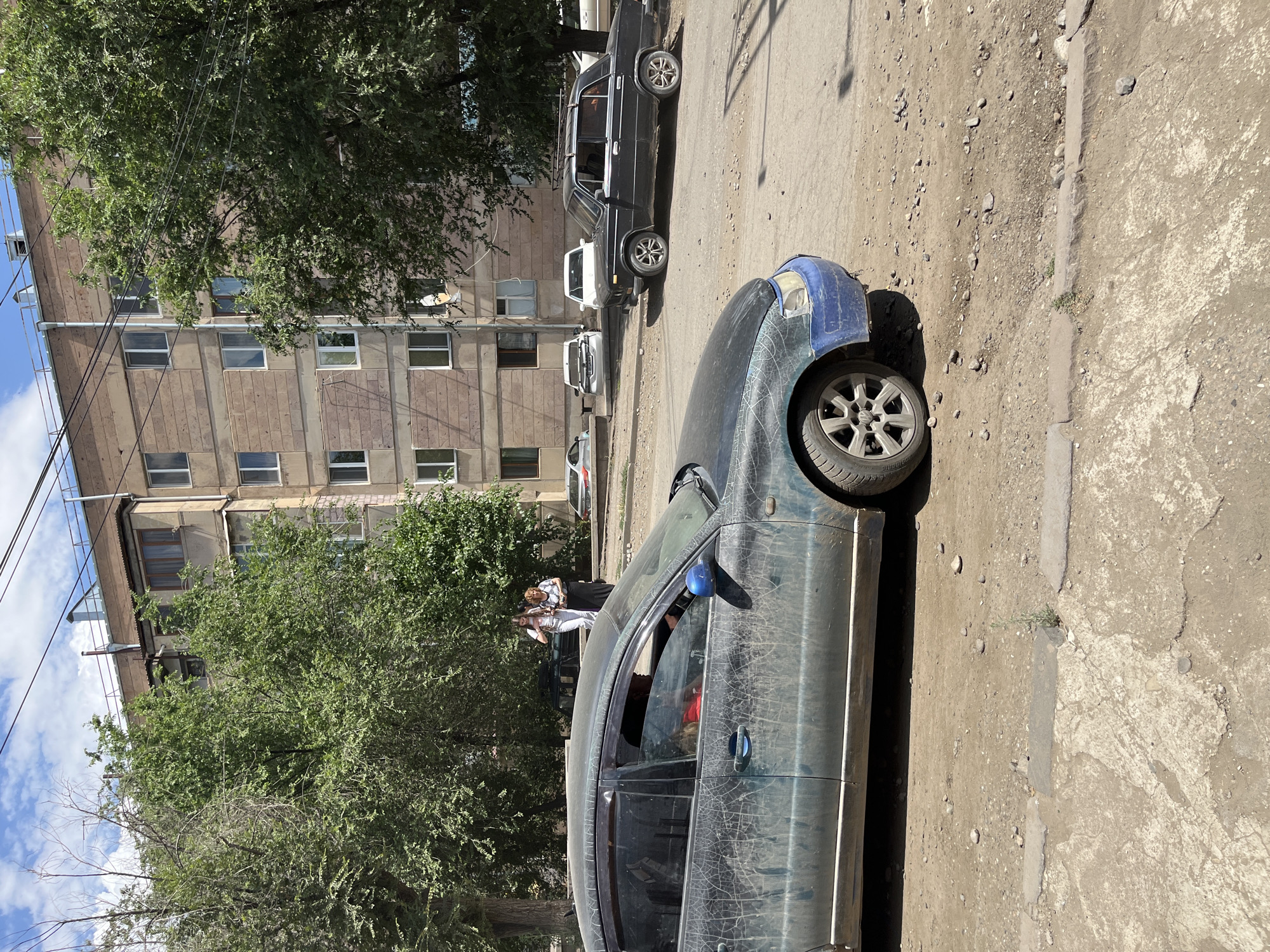 The image depicts a street scene in Gladzor, Armenia. In the foreground, a dark-colored, dirt-covered sedan is parked on a dusty, unpaved road. The car has a blue bumper. Behind it, a black sedan with a roof rack is also parked on the road. Further in the background, a multi-story apartment building stands, with trees in front of it. Two women are walking on the sidewalk in front of the building. The sky is partly cloudy. The environment appears to be an urban residential area with a mix of buildings and greenery.