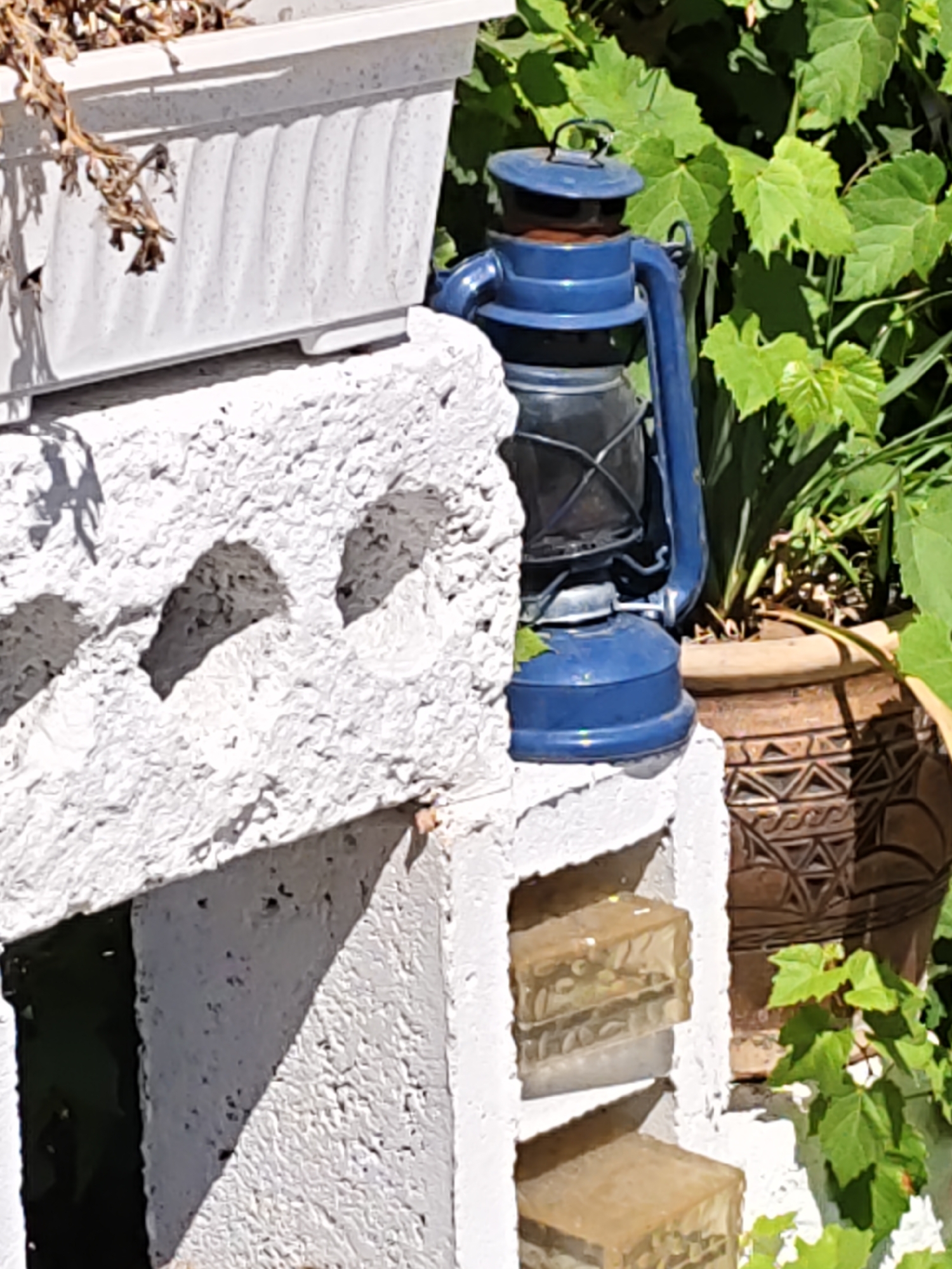 A close-up, eye-level shot captures a blue kerosene lantern placed on a white cinder block structure in a garden setting. The lantern, with its dark glass enclosure and metal handle, sits next to a white rectangular planter containing dried foliage. To the right of the lantern, a decorative terracotta pot with a patterned design is visible, partially obscured by lush green leaves of a vine. The cinder block structure features visible hollows and is partially filled with two translucent blocks containing what appear to be small pebbles or seeds. The scene suggests an outdoor decorative arrangement. The image is part of a dataset sourced from Rostov-on-Don, Russia, but no specific location within the city is identifiable from the visual content alone.