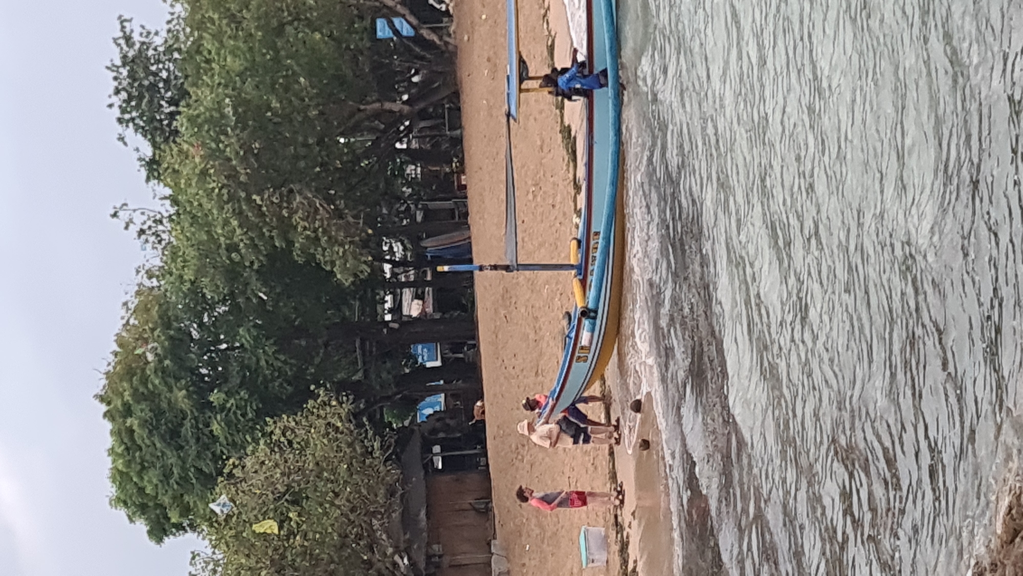 The image depicts a beach scene in Denpasar, Indonesia, with a traditional Indonesian fishing boat positioned near the shore. The boat is painted blue and yellow with some text on its side. Several individuals are visible on the sandy beach near the boat. On the left, two people are standing, one wearing a red and grey shirt and red shorts, and the other in a tan shirt and shorts. Closer to the boat, three people are gathered, two appearing to be interacting with the boat, and another person is inside the boat wearing a blue garment. The environment includes a sandy beach, trees in the background, and the ocean with gentle waves lapping at the shore. There are also some objects on the beach, including a white cooler and what appear to be coconuts. The sky is overcast.
