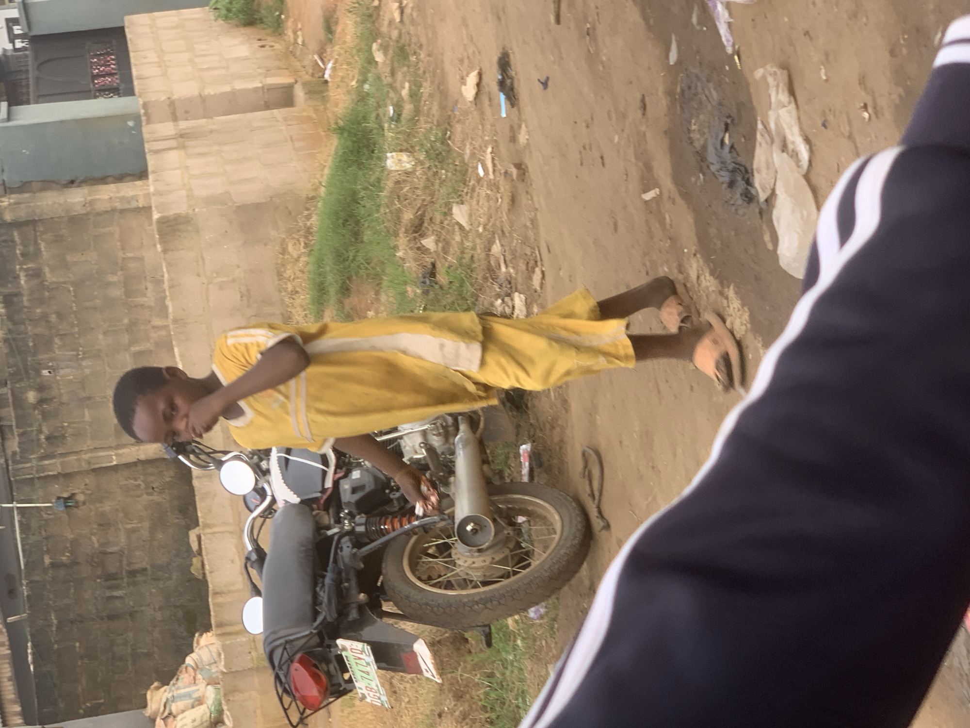 In the city of Shagamu, Nigeria, a young boy wearing a yellow outfit stands near a motorcycle on a dirt road. He is holding a white cloth to his nose. The motorcycle is black and white with a license plate that reads "DGB-742YD". In the background, a wall made of concrete blocks is visible, along with some sacks. The ground is covered in dirt, grass, and scattered debris, including plastic bags and other trash. The overall scene suggests an outdoor environment, possibly in a residential or semi-developed area of Shagamu.
