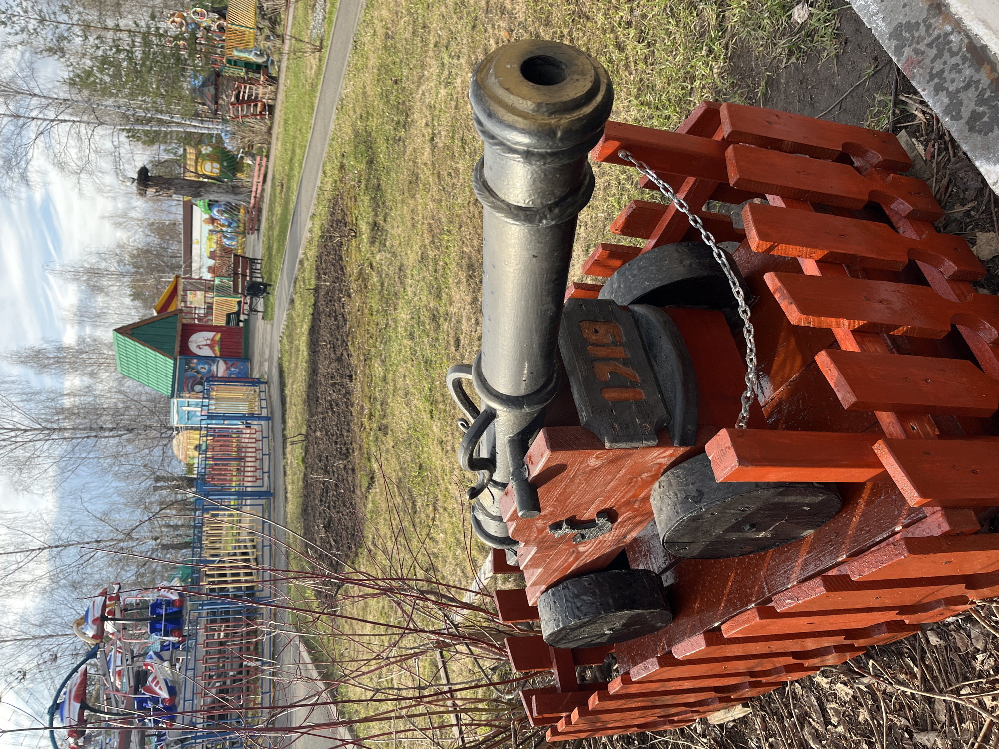 The image captures a decorative, dark-colored cannon with a prominent red wooden base, resembling a segmented track, in the foreground of a park or amusement area in Chaikovsky, Russia. A silver chain is draped from the cannon's barrel to its side. On the side of the cannon's base, the numbers "1719" are distinctly visible.
The scene unfolds under a clear sky with sparse, wispy clouds, suggesting a bright day, possibly morning or early afternoon, given the distinct shadows cast. The ground cover consists of patches of dormant, light-green grass and areas of bare, dark earth. Numerous bare trees with slender branches are scattered throughout the background, indicating either late winter or early spring.
The midground and background are populated with various colorful playground structures and amusement rides. These include a building with a vibrant green roof, fences with multi-colored slats, and what appears to be a small carousel or spinner ride featuring red, white, and blue elements. Other brightly colored play equipment, possibly a slide or climbing frame, are also visible. Paved pathways wind through the recreational space. No people are present in the image, giving the park a quiet and peaceful atmosphere, perhaps awaiting visitors.