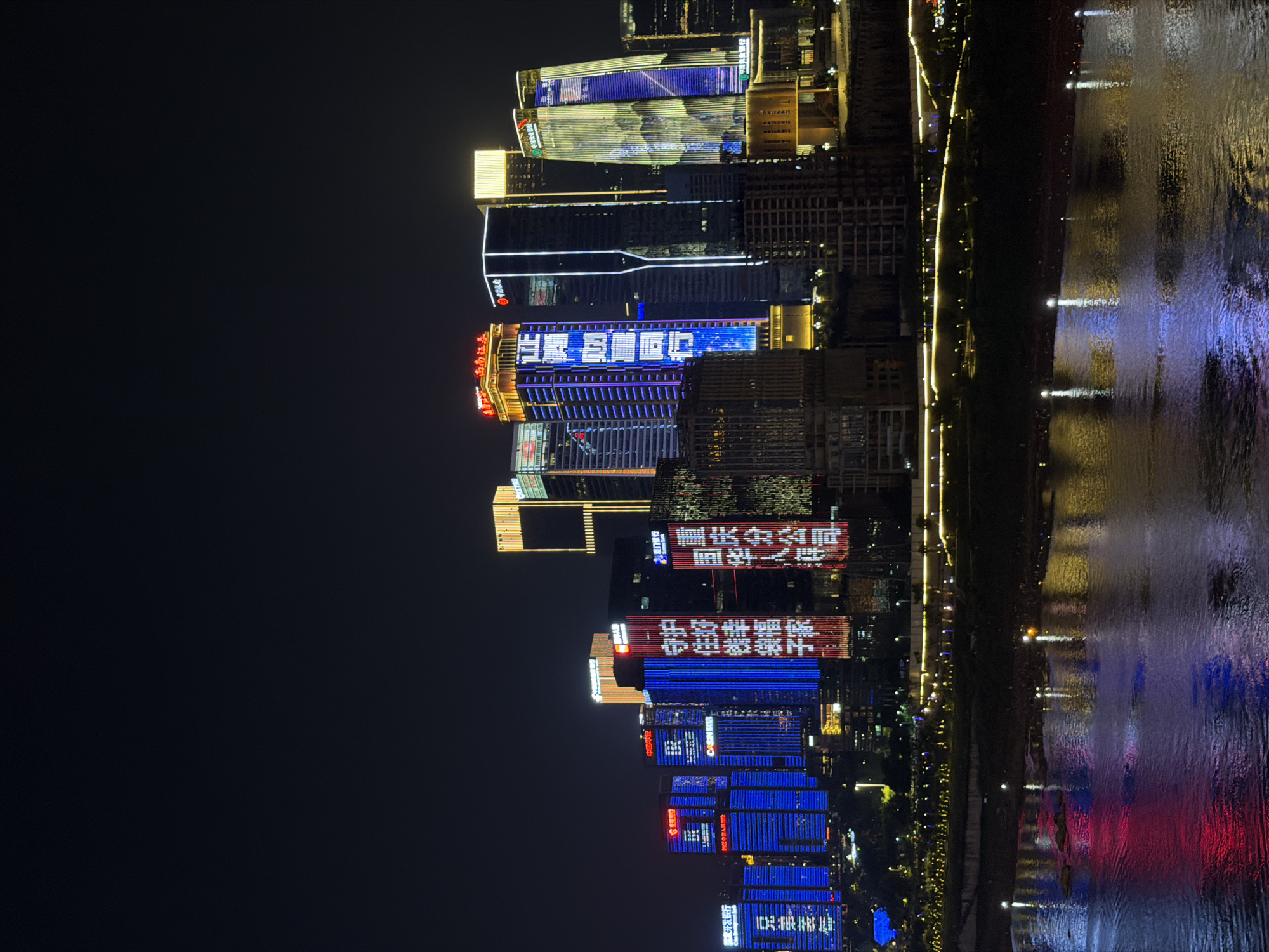 A nighttime cityscape features a river in the foreground reflecting the lights of a densely packed urban skyline. The buildings are illuminated with colorful lights, many displaying Chinese characters and logos. The most prominent buildings are modern skyscrapers with illuminated facades. On the left side, several blue-lit buildings show the characters "中国银行" (Bank of China). To their right, a skyscraper is lit with vertical blue text, and another building displays red characters that appear to say "守护伟大祖国" (Protect the Great Motherland). Further to the right, a building is illuminated with vertical yellow and blue lights, and another skyscraper has a yellow-lit top section. The river reflects the vibrant lights of the city, creating a colorful spectacle on its surface. The dark sky above contrasts with the illuminated city, suggesting a clear night. The scene captures the bustling urban environment of Yuzhong District, China.