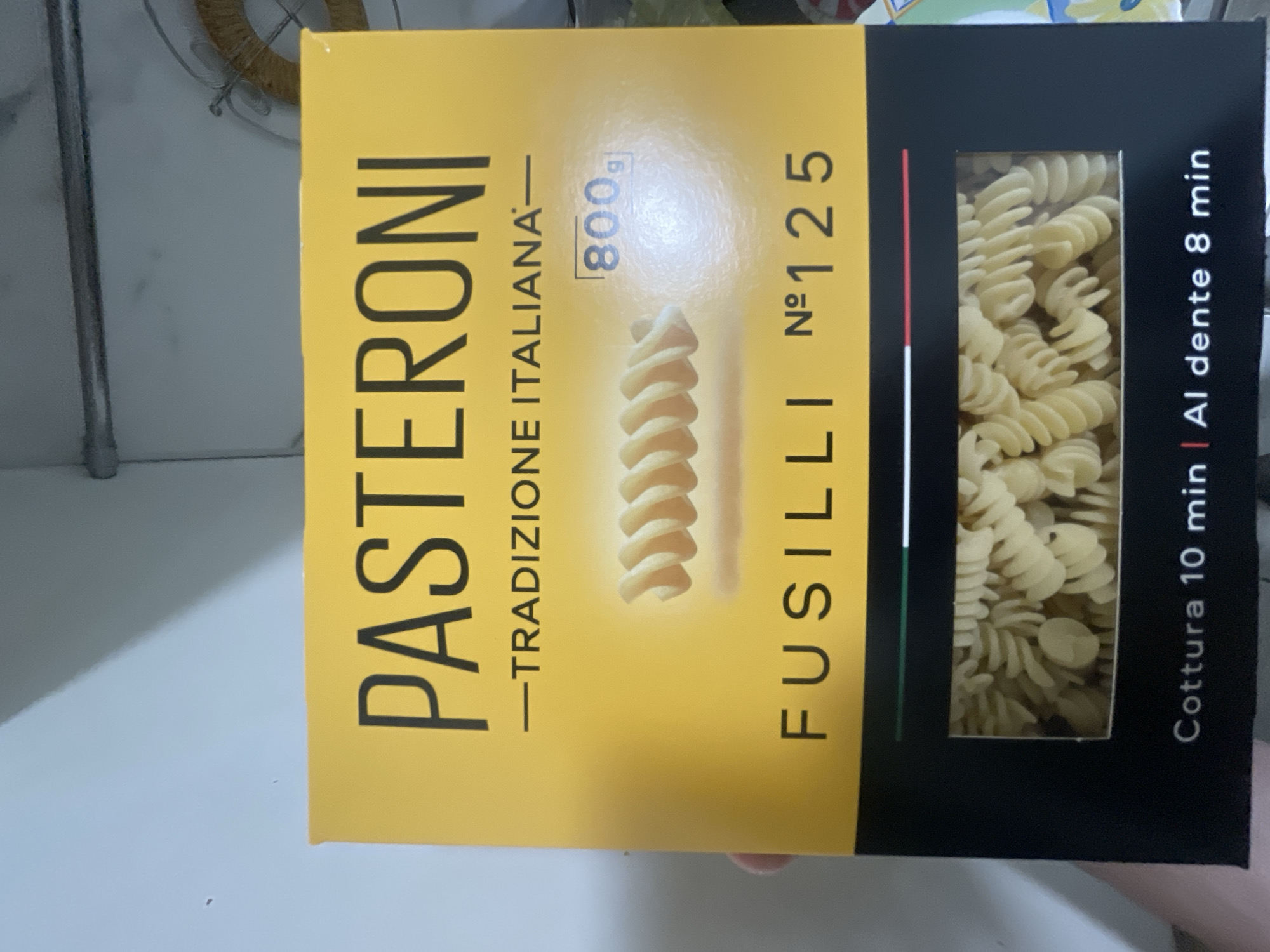 The image shows the front of a yellow and black box of Pasteroni Fusilli pasta. The brand name "PASTERONI" is prominently displayed in large black letters at the top of the box, below which is the text "TRADIZIONE ITALIANA". To the right of the pasta image, the weight is indicated as "800g". Below the pasta image, the text "FUSILLI N° 125" is visible. At the bottom of the box, the cooking instructions read "Cottura 10 min | Al dente 8 min". The box is partially held by a person's hand. The background is a plain light-colored wall and what appears to be part of a shelf or appliance. The image is taken indoors. There is no information in the image that relates to Bol'shoe Zaborod'e, Russia.