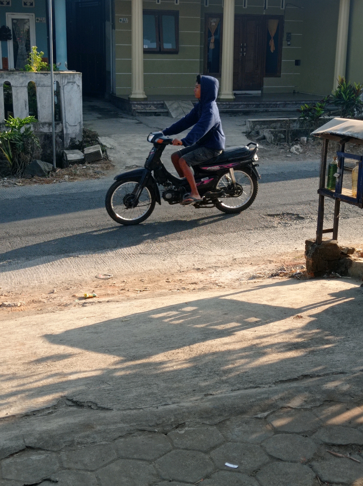 A person wearing a blue hoodie and shorts is riding a black motorcycle on a paved road. The person is facing to the left and appears to be looking forward. The motorcycle has a silver rim on the front wheel and a black rim on the rear wheel. The background shows a house with a light green exterior, white columns, and dark brown double doors. There are plants in front of the house. To the left of the motorcycle, there is a concrete wall and some greenery. The lighting suggests it is late afternoon, with long shadows cast on the road. The location is Purwosekar, Indonesia.