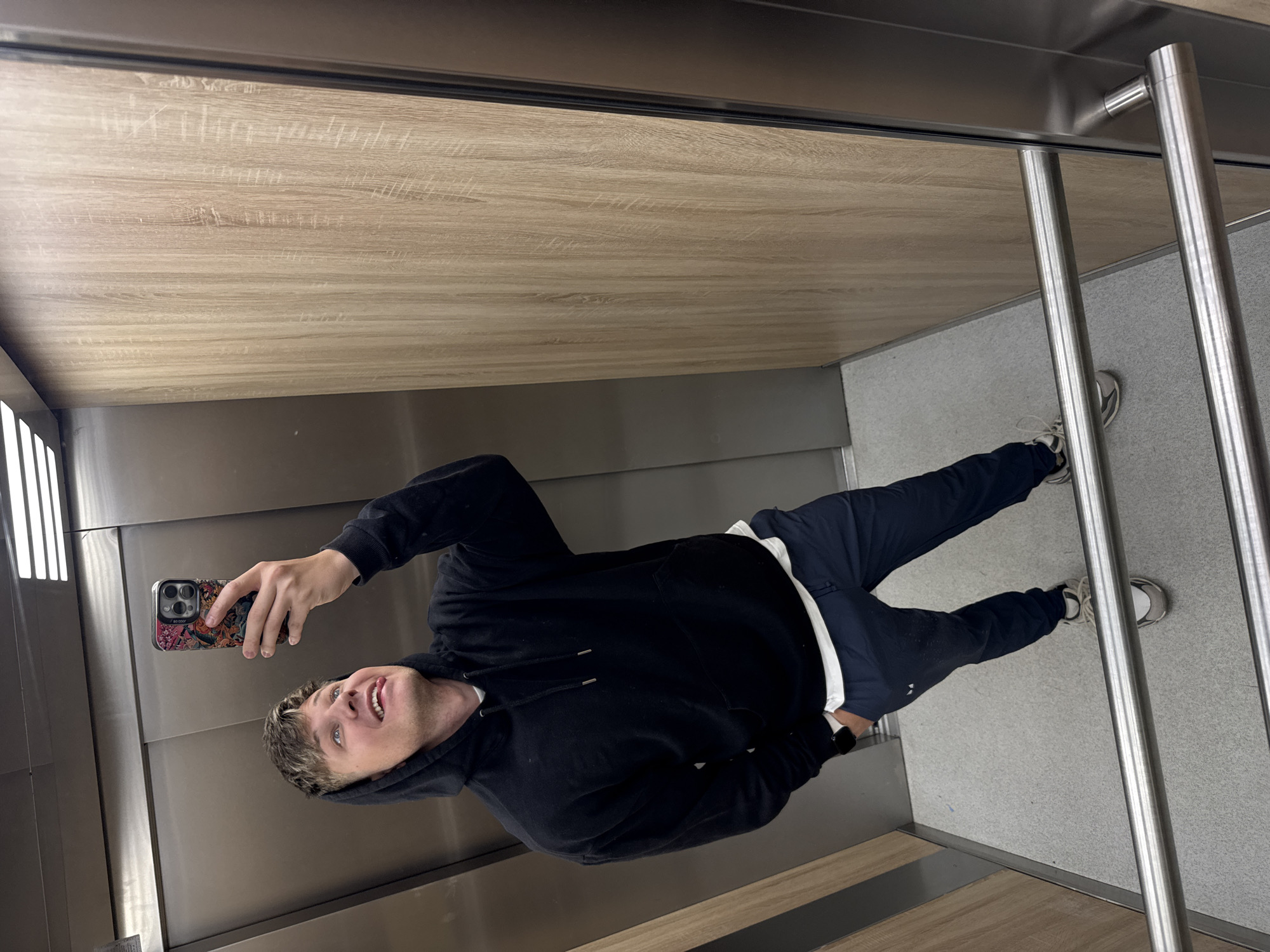 A young man is captured in a mirror selfie taken inside a modern elevator. He stands facing the mirror, holding his smartphone in his right hand to take the picture, with his left hand slightly obscured. He is smiling broadly with a playful expression, sticking his tongue out slightly.
The man is dressed casually in a black hooded sweatshirt (hoodie) over what appears to be a white undershirt or t-shirt, and dark blue sweatpants. White sneakers are visible on his feet, and a dark-colored watch is on his left wrist. His smartphone features a distinctive colorful case with what looks like comic book or anime-style imagery, and the text "BO COOP" is visible on it.
The elevator interior features a reflective surface that functions as a mirror, showing the man's full reflection. The top portion of the elevator's interior walls and ceiling has a light wood-grain patterned laminate, while the lower walls are a sleek metallic grey or silver. The floor is covered in a light grey, slightly textured material. On the right side of the image, two vertical metallic handrails are visible. In the background, to the left, there's a linear light fixture. A small, unreadable plaque or label is present on the lower left wall. The setting suggests a contemporary building in Plovdiv, Bulgaria.
