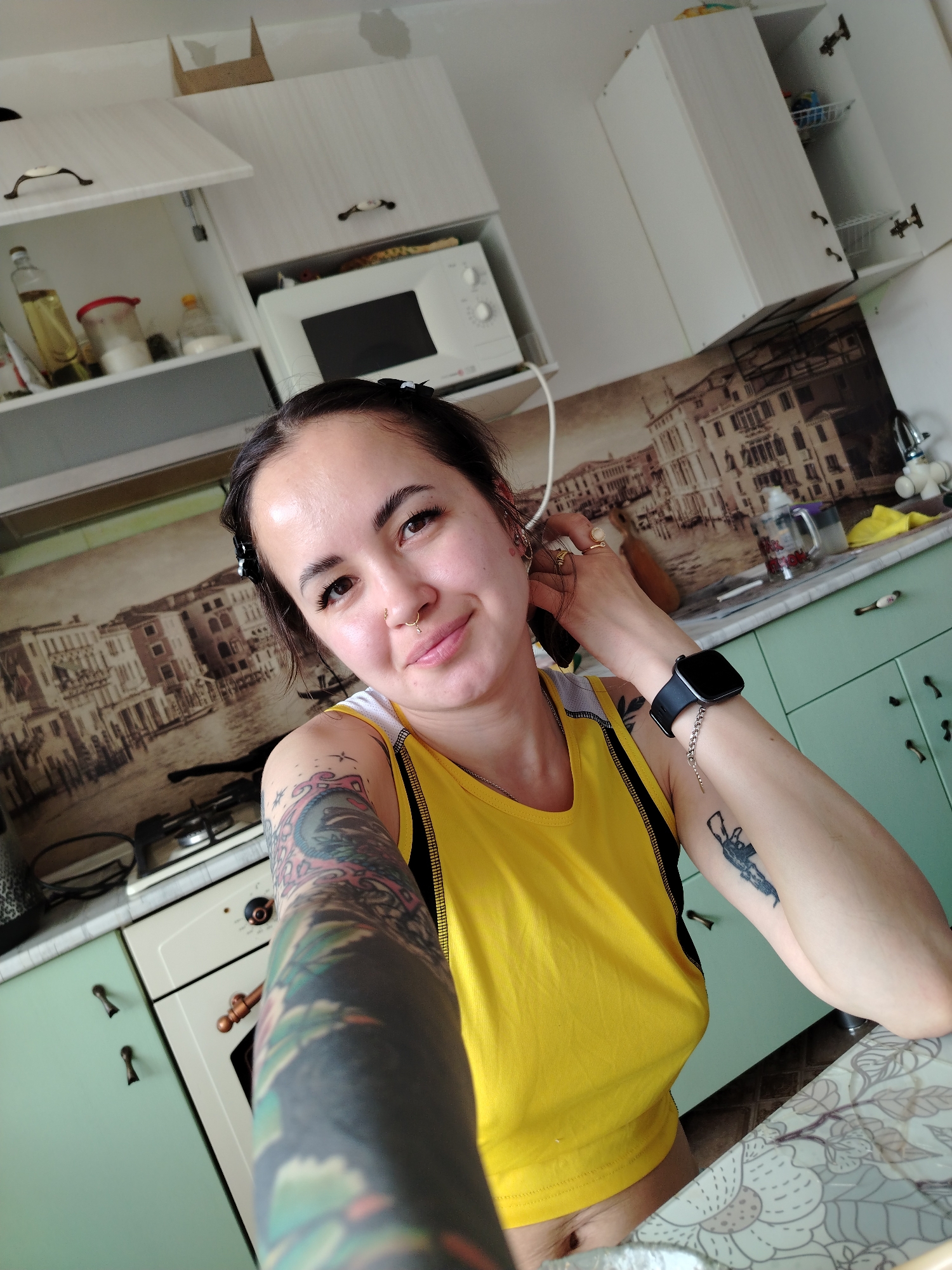 A woman takes a selfie in a kitchen. The woman has dark hair pulled back with a black hair accessory. She has fair skin and multiple piercings in her ears and nose. She wears a yellow tank top with black trim, exposing a small portion of her midriff. Her left arm, extending toward the camera, is covered in tattoos. She wears a smartwatch on her right wrist.
The kitchen features white cabinets, some with open doors revealing contents such as cooking oil and other items. A microwave is situated between the cabinets. The countertop behind the woman features a backdrop depicting a cityscape resembling Venice, Italy. On the countertop, there is a water bottle and a yellow cloth. The cabinets below the countertop are mint green. A white oven is visible to the left. The location of this scene is not in Noviy, Russia.