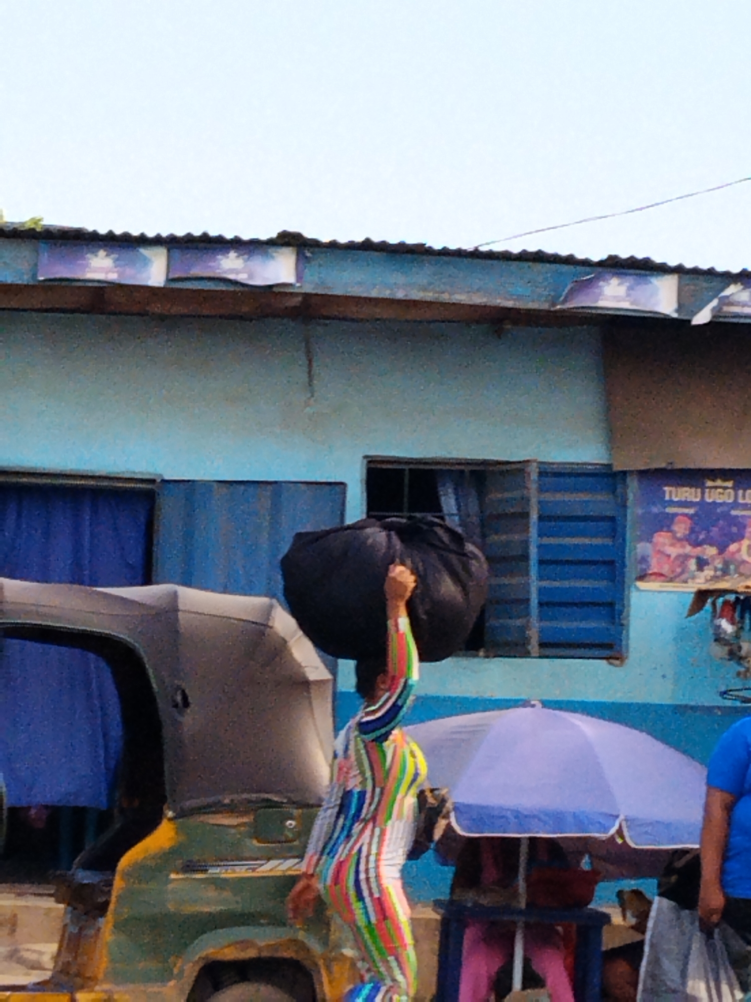 A woman in a colorful, striped jumpsuit is carrying a large black bag on her head in Aba, Nigeria. She is standing in front of a light blue building with a corrugated metal roof. A green and yellow three-wheeled vehicle, also known as a "keke," is parked to her left. A blue umbrella shades a stall with a person sitting beneath it to her right. A poster, with the visible text "TURU UGO LO...", is attached to the building.