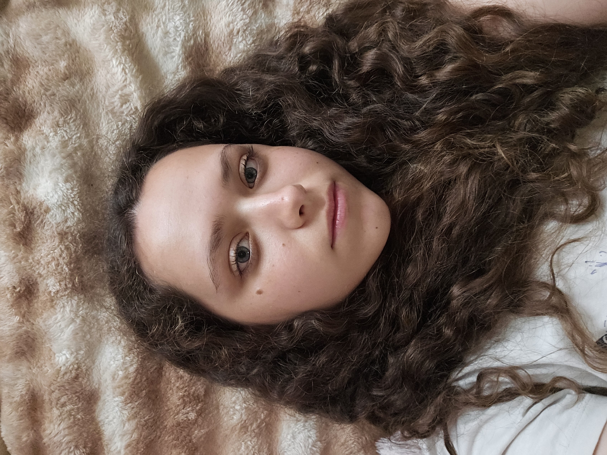 The media file contains a close-up, overhead shot of a young woman with long, curly brown hair, lying on a soft, textured, beige surface, possibly a blanket or bedspread. Her head is tilted, and she is looking towards the camera. Her eyes are light blue-green and open, and her lips are slightly parted, giving her a neutral to slightly curious expression. She has fair skin with a few freckles visible on her cheeks and nose. A mole is visible on her left cheek. The lighting suggests it may be daytime, as there is soft, diffused light illuminating her face. The background is out of focus, emphasizing the subject. A portion of a white garment, possibly a t-shirt, is visible on the right side of the frame, with faint blue markings that are illegible. The overall scene is intimate and personal, with the focus solely on the woman's face and hair. There is no discernible text in the image.