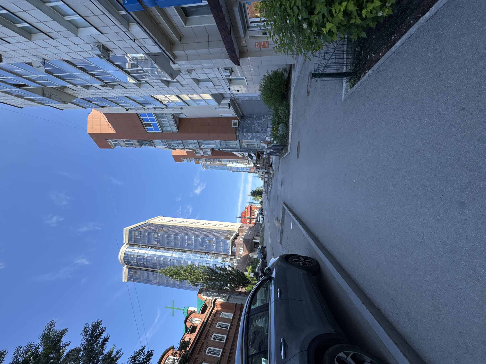 This is a daytime urban street scene in Perm, Russia, captured from a slightly elevated perspective looking down a street. The sky is bright blue with a few wispy white clouds, indicating clear, sunny weather.
On the left side of the street (viewer's left), a multi-story building with a light-colored facade and numerous reflective glass windows and balconies dominates the foreground. Sections of this building are clad in reddish-brown panels. Further down the street, a prominent, tall, modern skyscraper with a predominantly glass exterior and a distinctive curved upper section stands out against the sky. Below and slightly in front of the skyscraper, an older, reddish-brown brick building with white window frames and a green cross on its roof suggests a church or religious institution.
On the right side of the street, a dark grey SUV-like car is parked parallel to a concrete curb and sidewalk. A stretch of green bushes and a metal fence line the sidewalk, separating it from the roadway. Beyond these, another building, less distinct, is partially visible.
The street itself is paved with asphalt and extends into the distance, with more buildings partially visible further along. No people are visible, and no specific activity is taking place, presenting a static snapshot of an urban environment. A few small signs are visible on buildings, but no legible text can be discerned. The scene showcases a blend of older brick architecture and modern glass structures.