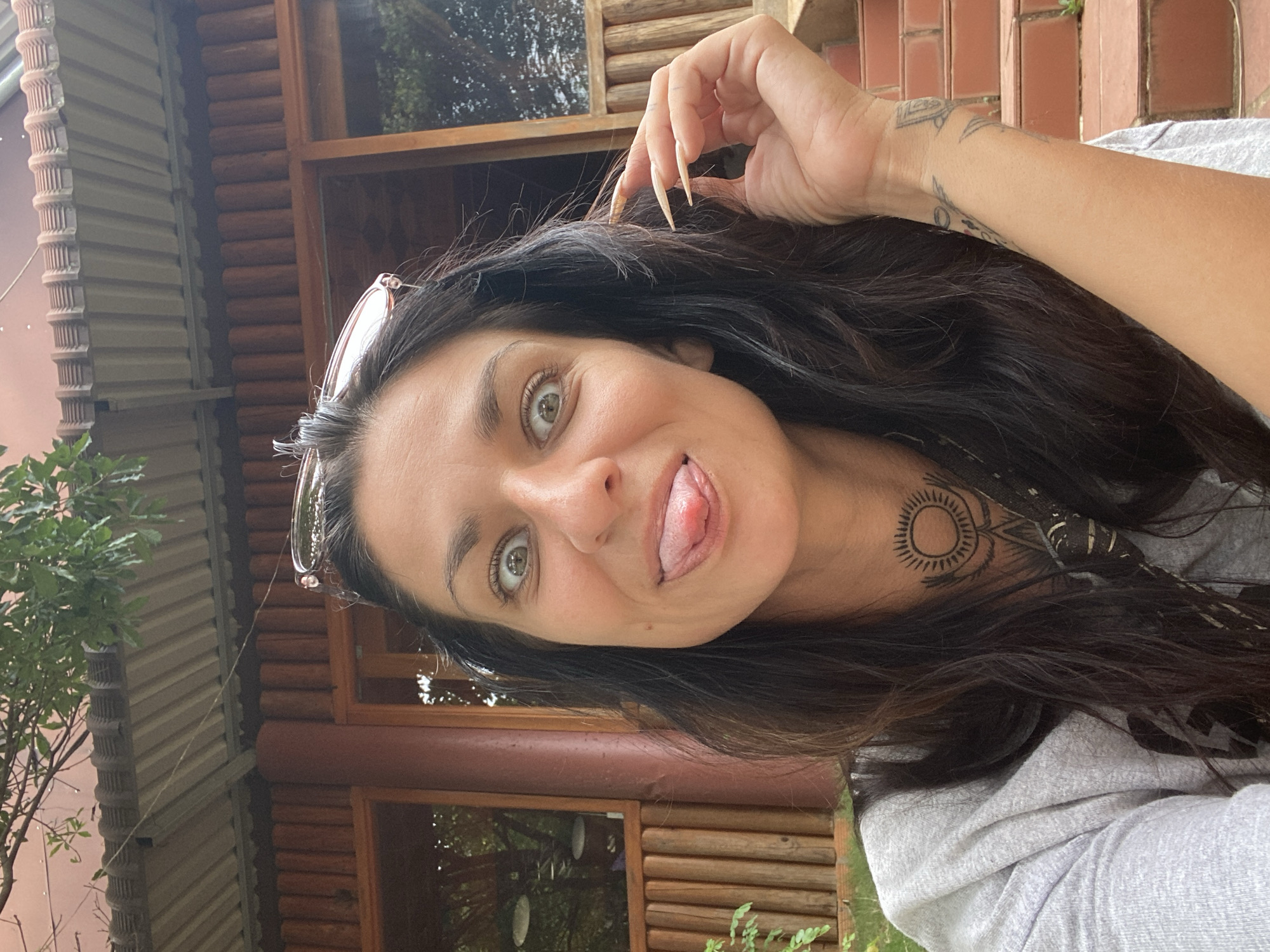 A medium shot captures a woman with dark hair and a playful expression, sticking her tongue out at the camera. She is wearing a grey t-shirt and sunglasses perched on her head. A tattoo is visible on her neck. The background features a rustic building with wooden beams and a corrugated metal roof, suggesting an outdoor setting. There are also some steps made of red brick visible in the background. The environment appears to be natural, with greenery visible around the building. The location is not specified as Thôn 2, Vietnam, as this information is not derivable from the image.