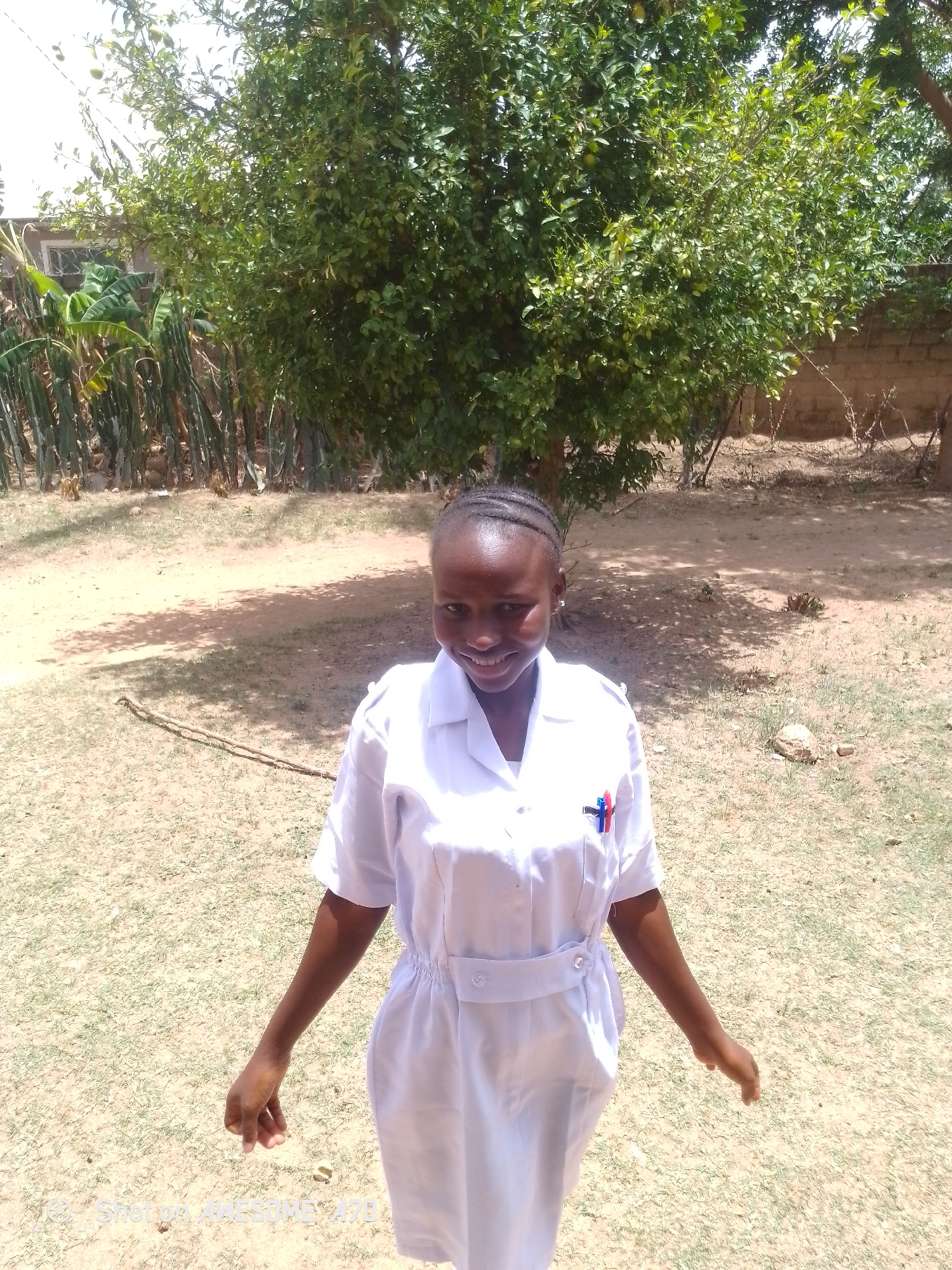 A young girl stands in a grassy area in an unspecified location within the city of Unknown, Unknown. She is wearing a white uniform dress with a small red and blue emblem on the chest. Behind her is a large, leafy tree providing shade, and in the background are elements suggesting a residential or rural setting, including what appears to be a brick wall and some plants. The ground is mostly bare with scattered grass.