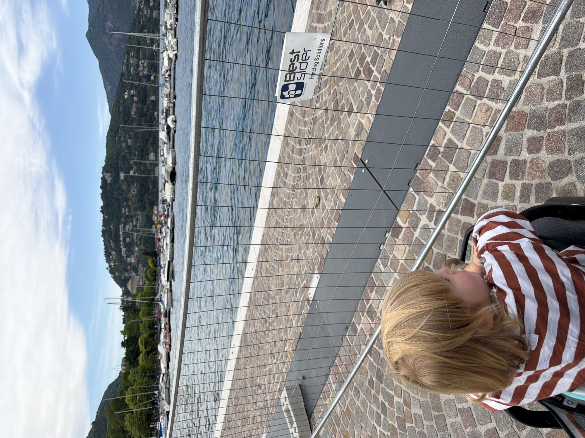 A young child with blonde hair, wearing a striped shirt, is seated in a stroller on a paved walkway. A wire fence with a "Best Sider" sign separates the walkway from the water. Behind the fence is a view of water, boats, and a mountain covered in trees and buildings. The scene is in the city of Como, Italy.