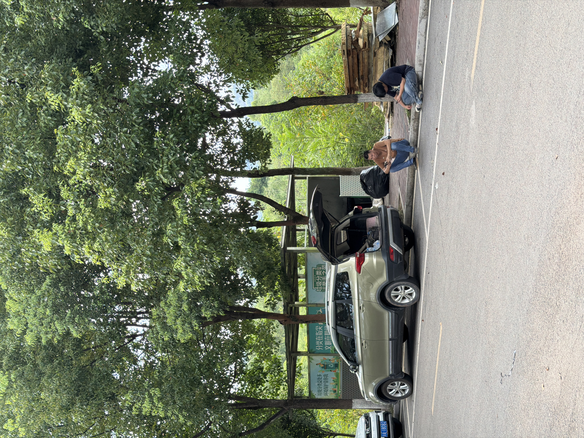 Two men are seen squatting on the side of a road in Jiangjin District, Chongqing City, China, with their backs to a parked car. Both men are looking down at their mobile phones. The man on the left is wearing a tan t-shirt and blue jeans, while the man on the right is wearing a dark t-shirt and blue jeans. Behind them, a gold SUV is parked with its trunk open, revealing bags inside. The scene is set against a backdrop of lush green trees and vegetation, with a small roadside shelter visible further in the background. To the right of the men, a stack of lumber is piled up next to some discarded building materials. The road in the foreground is asphalt, marked with yellow lines.