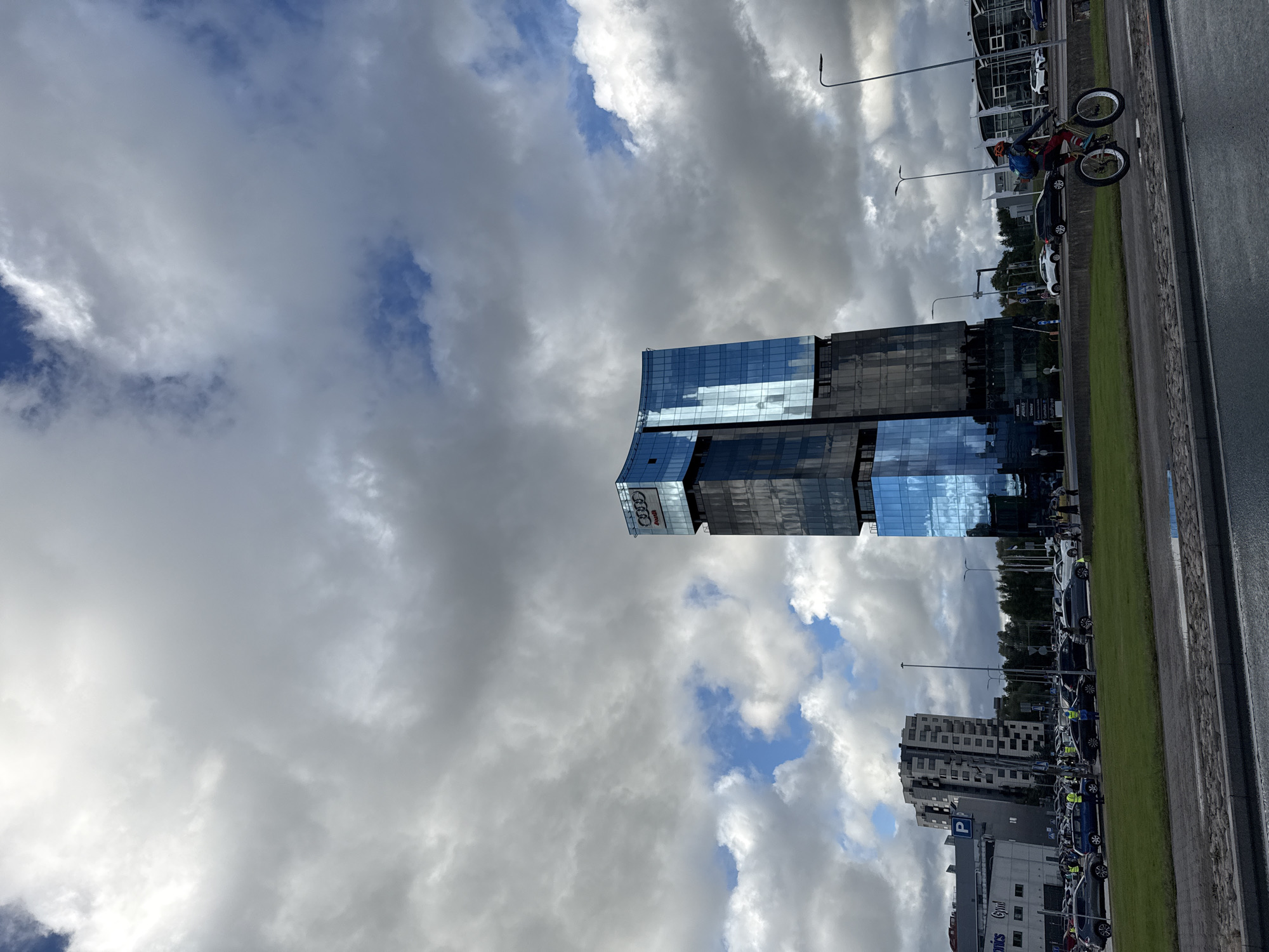 The image captures a wide shot of a street scene in Tallinn, Estonia, dominated by a modern, glass-fronted skyscraper featuring the Audi logo prominently at its peak. The skyscraper's reflective facade mirrors the cloudy sky, which is a mix of dramatic grey storm clouds and patches of blue sky. In the foreground, a person on a fat-tire bicycle is in motion, their back to the camera, wearing a backpack. The bicycle has large, knobby tires, suggesting it is built for rough terrain or perhaps snow. The cyclist is on a paved road, with a grassy median strip to their left. Beyond the grassy strip is a parking lot filled with numerous cars, many of them appearing to be white or silver. Several individuals, some wearing yellow vests, are visible in the parking lot, suggesting an organized event or a busy day at the Audi dealership. To the left of the Audi building, another, squatter, grey building with many windows is visible, along with signs for "Gym" and "Auto". The overall environment appears to be a commercial or business district, with the Audi building serving as a landmark. The weather suggests a dynamic, possibly overcast day with intermittent sunshine.