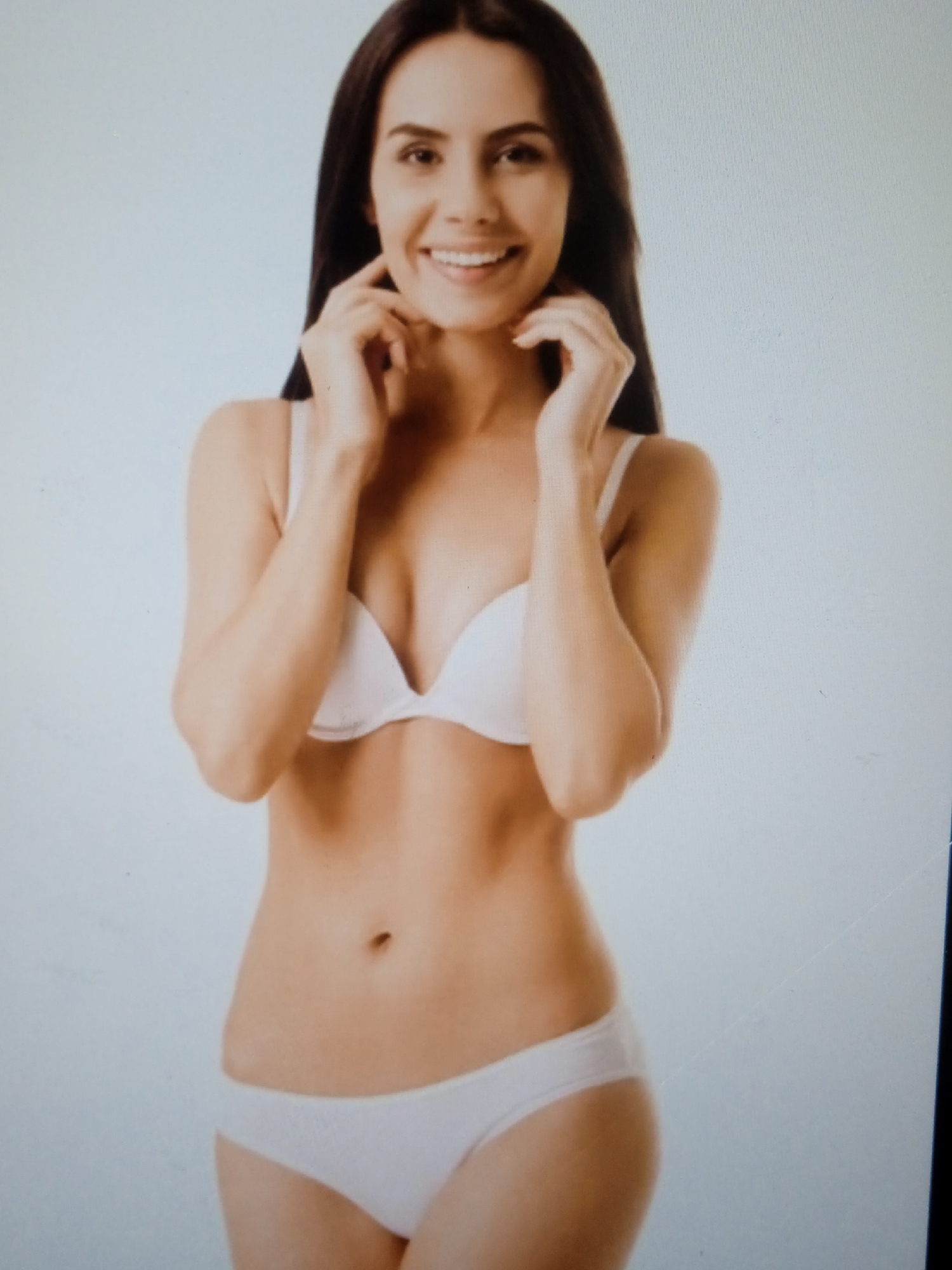 The image shows a full-body studio portrait of a young woman with long, dark hair and a light complexion. She is wearing a white bra and white underwear. She is smiling and has her hands near her face. The background is a plain, light blue. There is no indication of the image being taken in Chernivtsi, Ukraine.