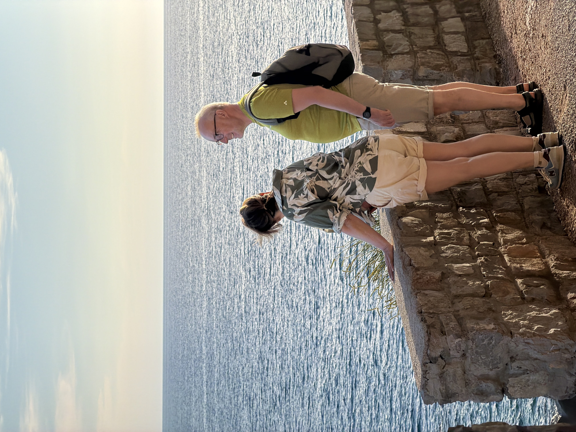 Two people, a man and a woman, stand near a stone wall overlooking the sea. The man, on the right, wears glasses, a lime green t-shirt, khaki shorts, and black sandals. He carries a gray backpack. The woman, on the left, faces away from the camera, wearing a patterned shirt and light-colored shorts. She leans on the wall, her hand resting on the stone. The sea stretches out to the horizon under a pale blue sky with wispy clouds. The scene appears to be in Petrovac centar, Montenegro, likely along the promenade or a scenic overlook.