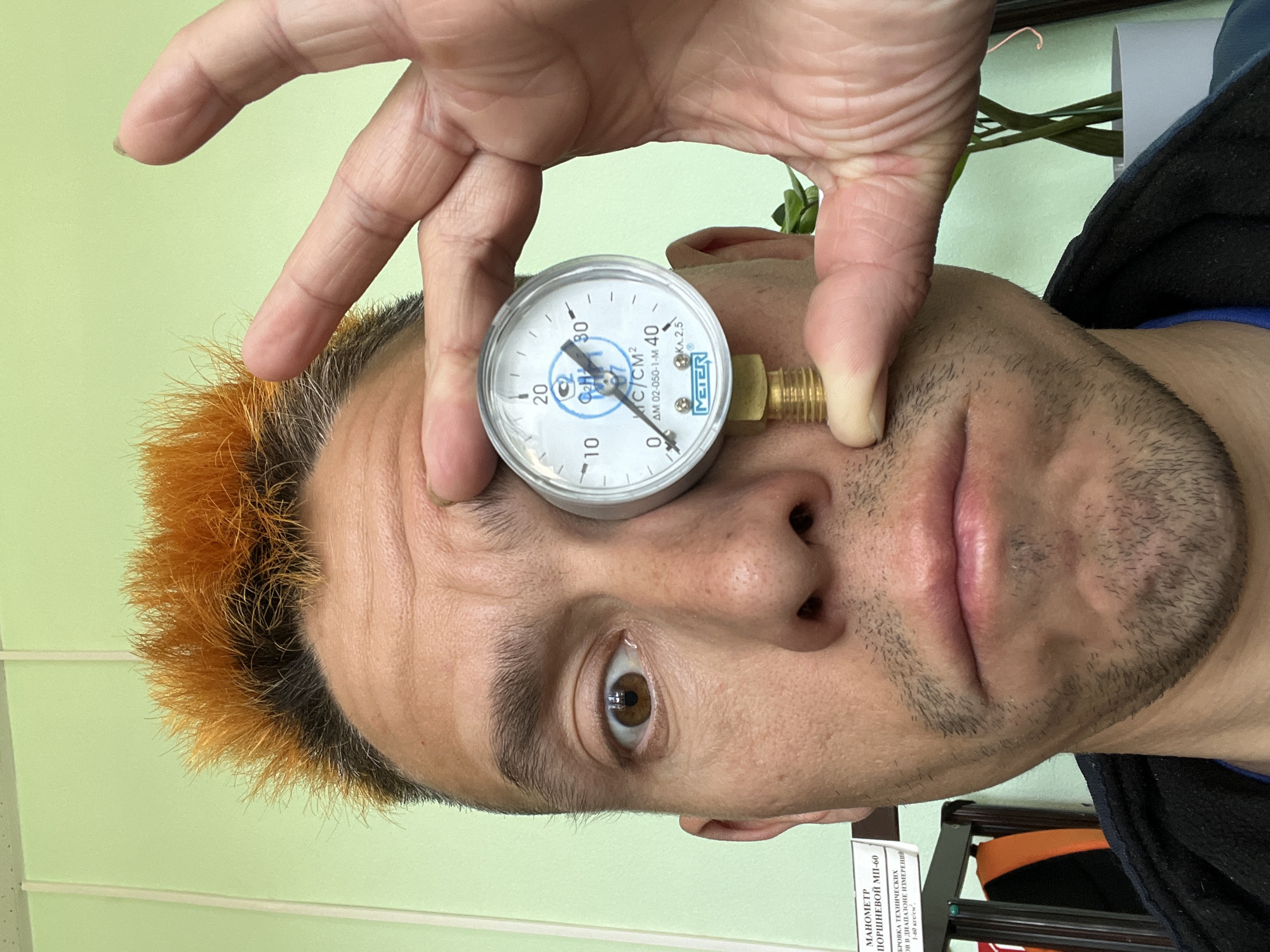 A man with orange-tipped hair is holding a pressure gauge to his eye. The gauge is white with black numerals and a brass connector. The man's right eye is visible behind the gauge. The background includes a light green wall and, out of focus, shelves and a plant. A sign with Russian text mentioning "Chaikovsky" and "MAHOMETP" (Manometer) is partially visible in the lower right corner. The photograph appears to be taken indoors.