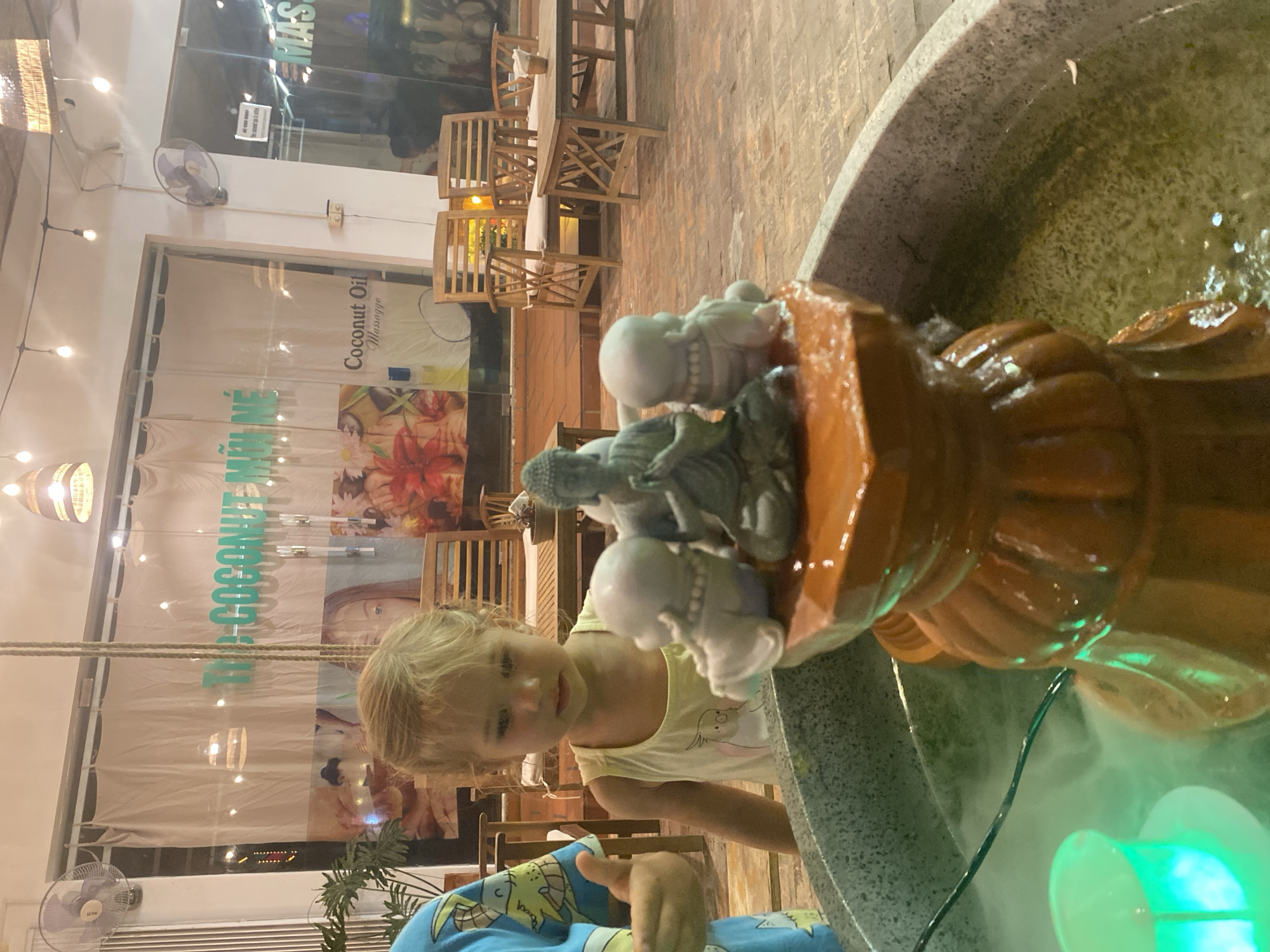 In Mũi Né, Vietnam, a young Caucasian girl with blonde hair, wearing a yellow tank top, stands near a water fountain. The fountain features a tiered, reddish-orange base adorned with small statues of a meditating Buddha and two accompanying figures. A green light source is visible submerged in the fountain's water, creating a misty effect. Behind the girl, a large window displays a sign with text that appears to be in Vietnamese, alongside images of flowers and text that reads "Coconut Oil Massage." Wooden chairs and tables are visible in the background, suggesting an outdoor seating area, possibly at a cafe or spa. The lighting indicates it is evening.