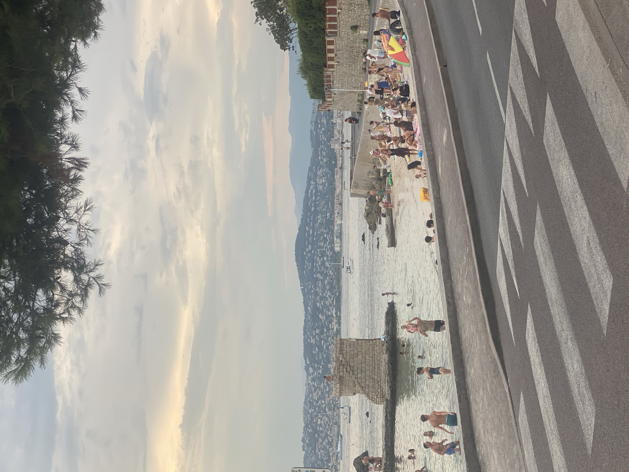The image depicts a lively scene at a beach in Antibes, France, likely during the summer season. In the foreground, a street with a prominent pedestrian crossing is visible, suggesting proximity to a public area. The beach itself is populated with numerous individuals enjoying the water and sand. Some are swimming, while others are sunbathing or relaxing on towels. Children are present, indicating a family-friendly atmosphere.
Further out in the water, a small, weathered stone structure stands as a unique landmark, possibly an old watchtower or part of a historical defense system. People are seen interacting around this structure, with one individual perched on top. Sailboats and other vessels dot the sea in the background, adding to the picturesque coastal scenery.
The coastline stretches into the distance, lined with buildings that cascade up the hillsides, characteristic of a Mediterranean town. The sky is partly cloudy, with soft light suggesting either early morning or late afternoon. The overall impression is one of a bustling, yet relaxed, beach environment in a beautiful coastal city.