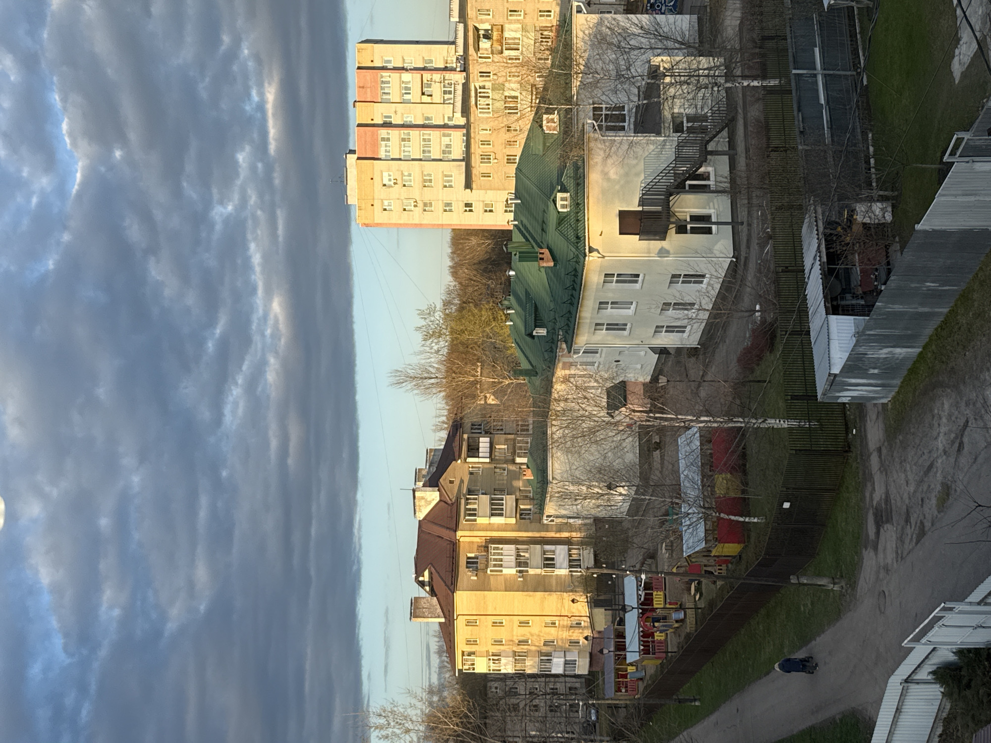 The image displays an urban residential scene, likely in Rusanovo, Russia, under a partly cloudy sky during either late afternoon or early morning. The perspective is elevated, looking down upon a series of buildings, paths, and green spaces.
Dominating the background are several tall, multi-story apartment buildings with light-colored facades (beige, off-white, and light orange) and numerous windows, some showing air conditioning units. In front of these, there are smaller multi-story buildings, one prominent structure featuring a green, sloped roof, and others with flat roofs, one of which includes an external black metal staircase. Utility sheds and structures with corrugated metal roofs are also scattered throughout.
The ground level features paved paths or roads, surrounded by patches of grass and bare trees, indicating a colder season like late autumn, winter, or early spring. Various fences, including wooden slat and metal varieties, divide the different areas. A colorful playground structure with red, yellow, and blue elements is partially visible behind a fence on the right. A single street lamp post stands near the center.
The sky is filled with textured clouds, ranging from dark grey to bright white, with clear patches of blue sky. A bright spot in the upper left corner suggests the sun's position or a very bright cloud. The low-angle sunlight creates distinct shadows and highlights on the buildings' facades.
A solitary person, dressed in a dark coat, is seen walking away from the viewer on a paved path in the lower right section of the image. A blurry blue circular logo is visible on the side of a white building in the mid-ground, but no specific text is discernible. The architecture and general setting align with typical residential areas in post-Soviet countries. The scene conveys a sense of quiet, everyday life.
