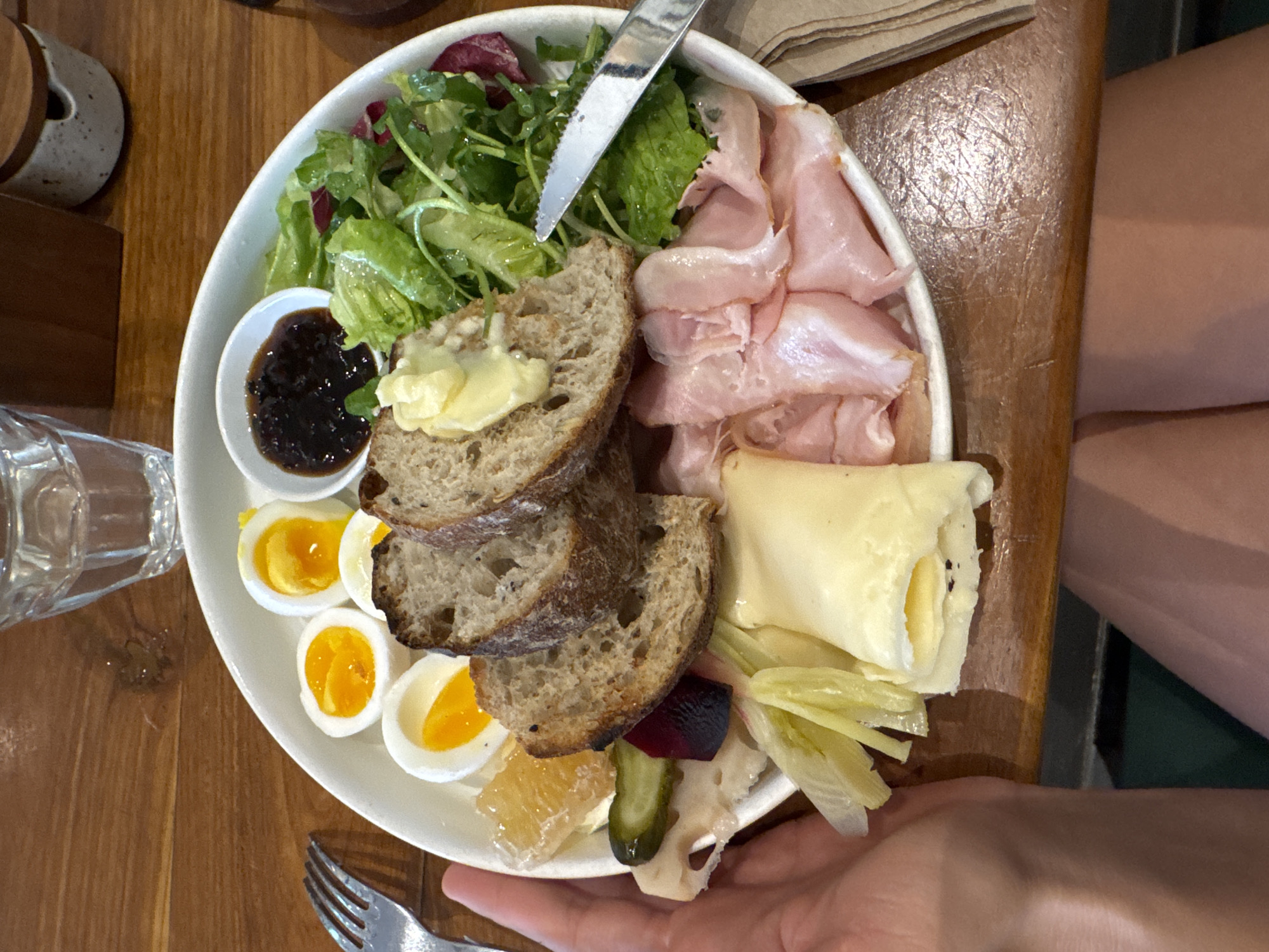 A top-down shot depicts a white plate filled with a variety of food items on a wooden table. The plate contains mixed greens, slices of ham, hard-boiled eggs cut in half, slices of bread with butter, a small bowl of dark jam, rolled cheese, and pickled vegetables. A silver knife is placed on top of the greens. A clear glass of water is located to the left of the plate, and a folded napkin is on the right. A fork is visible near the bottom left edge of the plate, and a hand is seen holding the plate from the bottom right. A leg of the person holding the plate is visible on the right side of the image. The table is made of wood, and a small ceramic container is visible in the upper left corner. The image was taken in the city of Ungasan, Indonesia.