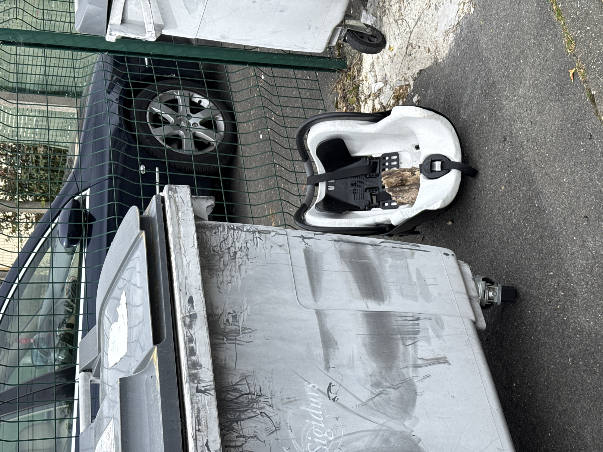 The image shows a street scene, likely in Domont, France based on the "Sigidurs" inscription on the garbage bin, as Sigidurs is a waste management authority in that region. A gray garbage bin is prominently featured, with visible wear and tear, and appears to be next to a green metal fence. A black car is partially visible through the fence. Next to the garbage bin and on the pavement, there is a discarded white baby car seat with black straps. The surrounding environment is a paved surface, likely a sidewalk or street.