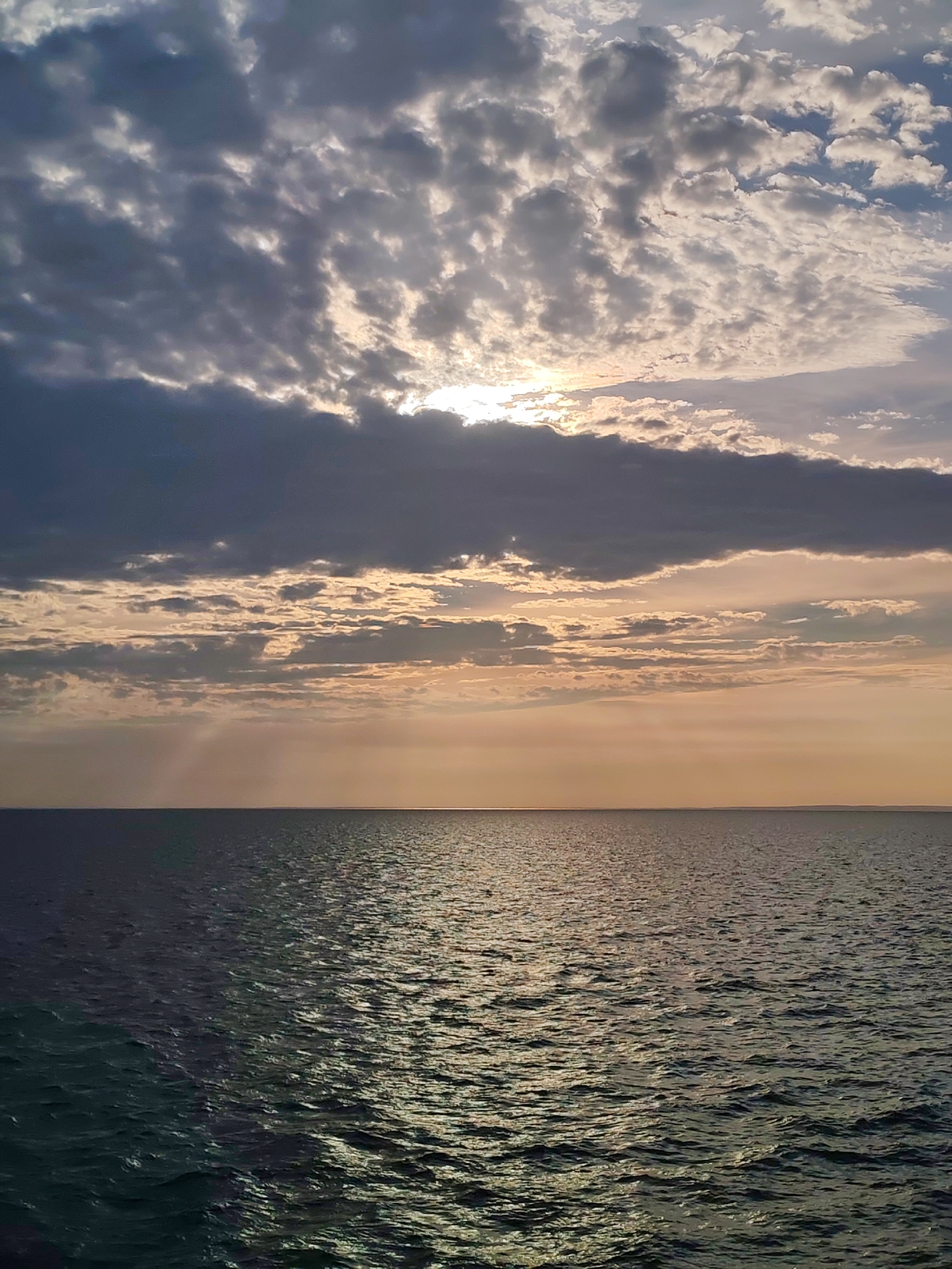 A wide shot captures a body of water and the sky above. The water fills the bottom half of the frame, appearing dark and somewhat choppy, reflecting the light from the sky. The horizon line is sharp and straight, separating the water from the sky. The sky dominates the upper half of the image, filled with a dramatic arrangement of clouds. Towards the left and center, dark, heavy clouds are interspersed with patches of lighter, brighter clouds, suggesting the sun is partially obscured. The sun itself is visible as a bright spot peeking through the clouds, casting a warm, golden light. The clouds are textured and varied, creating a sense of depth and movement. The overall impression is of a dramatic sky over a vast expanse of water, likely during sunrise or sunset. The location is Ozernoe Ust'e, Russia.