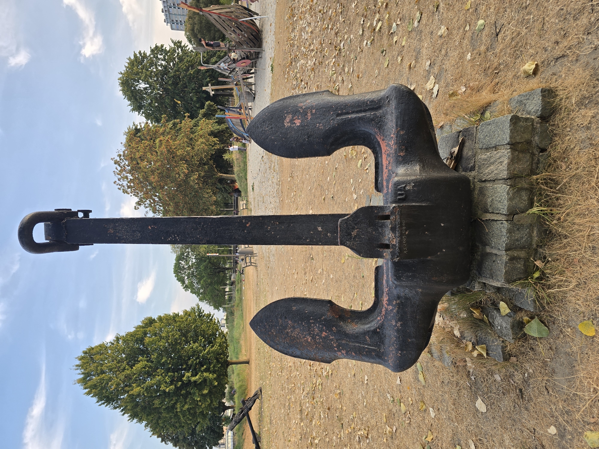 An antique, black, cast-iron anchor with red accents rests on a bed of grey stones in the foreground. The anchor is centrally located and commands the viewer's attention. Behind the anchor, a sandy area is strewn with dry leaves. In the mid-ground, several trees with green and autumnal foliage stand tall, casting shadows on the ground. To the right, a wooden playground structure resembling a ship is visible, with a few children playing on it. A couple of people are seated on a bench in the distance. In the far background, a modern, multi-story building can be seen, indicating an urban setting. The sky above is a clear blue with scattered white clouds. This scene appears to be at a park or recreational area along the waterfront in the city of Antwerp, Belgium.