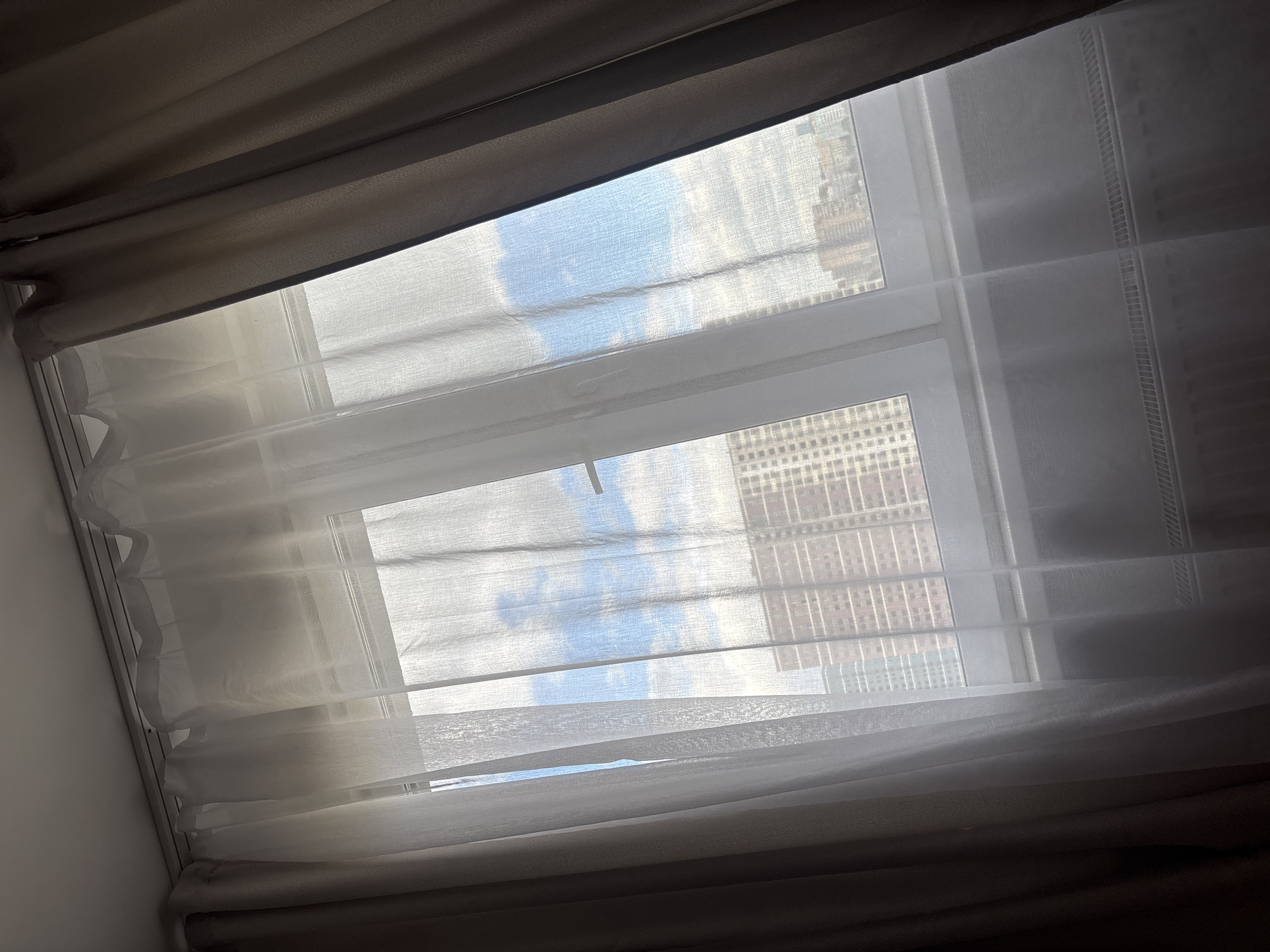 The image shows a view from inside a room, looking out through a window. The window is partially covered by two layers of curtains or blinds. The outer layer is a heavier, darker fabric, while the inner layer is a sheer, white material. Through the window, one can see a cityscape under a cloudy, blue sky. The buildings appear to be high-rise apartments or office buildings. The precise location within the city is not identifiable from the image.