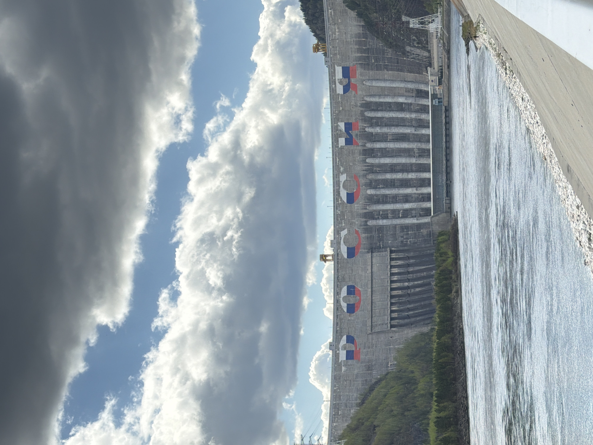 The image shows a side view of the Sayano-Shushenskaya Dam on the Yenisey River in Cheremushki, Russia. The dam is made of concrete and has large letters in the Russian flag colors. Water is flowing over the dam. The sky is partly cloudy. There is vegetation on either side of the dam.