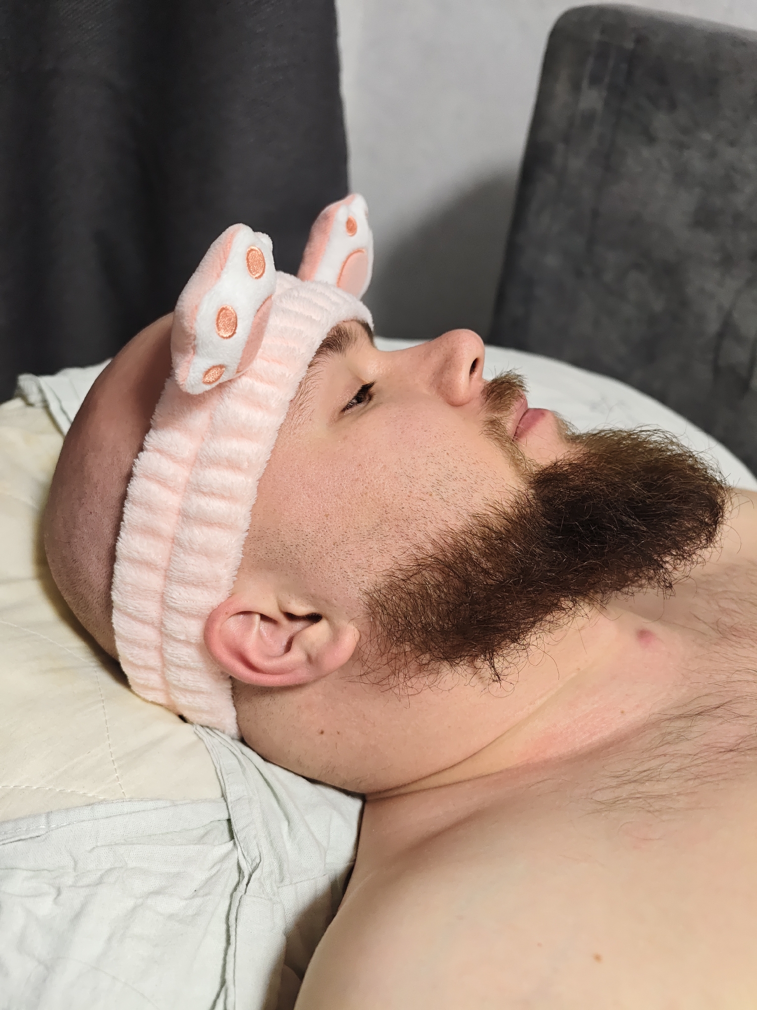A man with a bald head and a full brown beard and mustache is depicted lying on a light-colored pillow. He is wearing a light pink, fluffy headband adorned with two prominent bunny ears, which have lighter pink interiors and small orange-brown circular patterns resembling paw prints. His head is turned slightly to the right, and his gaze is directed upwards, suggesting a relaxed state.
The setting appears to be indoors, possibly a bedroom or living room, indicated by the pillow and bedding. In the background, a dark grey curtain is visible on the left, and a dark grey upholstered chair or piece of furniture is present on the right. The man's upper torso is partially visible, showing fair skin and some body hair. The lighting is soft and ambient, with no specific indicators of the time of day. The overall impression is one of calm and rest. No text is visible in the image. The implied location context is Chaikovsky, Russia.