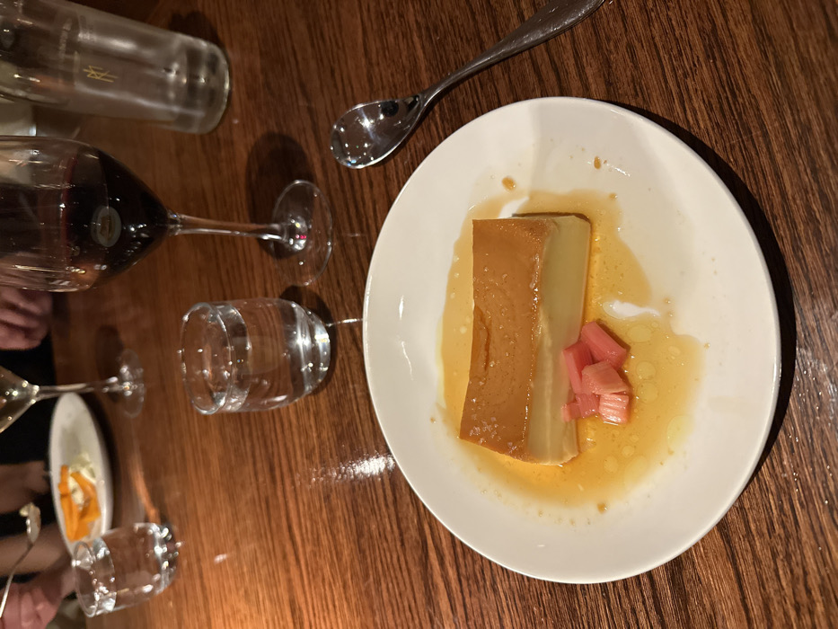 The image displays a close-up view of a dessert plate on a dark brown wooden table, indicative of a restaurant setting. In the foreground, a white ceramic plate holds a rectangular portion of flan or crème caramel, light yellow in color with a darker golden-brown top layer. It rests in a shallow pool of translucent amber liquid, presumably caramel sauce. Three small, pinkish-red pieces of what appear to be pickled rhubarb are positioned alongside the flan. To the right of the plate lies a silver spoon.
Various drinking vessels are arranged on the table around the main subject. Directly behind the dessert, a small clear glass contains water. Further back and to the right, a stemmed glass holds dark red liquid, identified as red wine. To the left, a partial view of another stemmed glass is visible, containing a lighter-colored liquid, possibly white wine. A clear glass bottle, partially visible on the far right, appears to hold water. In the upper left background, a person's hand holding a spoon is visible, interacting with a partially obscured white plate bearing orange food items. Another hand is vaguely discernible in the upper right. The lighting is dim, suggesting an intimate or evening dining atmosphere. The specific location within the city of London, United Kingdom, is not discernable from the visual information provided.