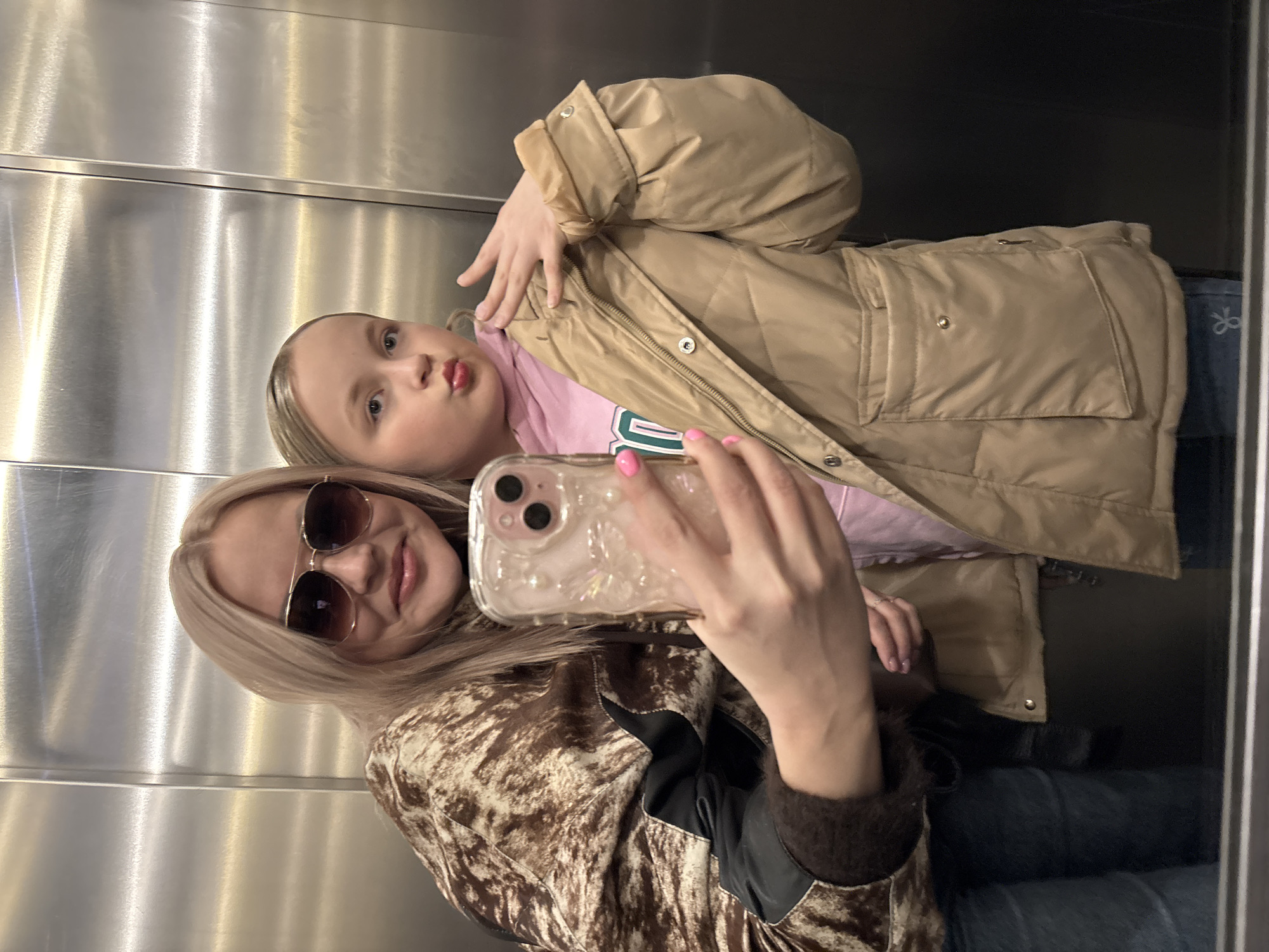 The image shows two fair-skinned individuals, one adult and one child, taking a selfie in what appears to be an elevator. The adult is wearing sunglasses and a patterned jacket with black leather accents on the shoulders and sleeves. The child, positioned slightly behind the adult, is wearing a light brown quilted jacket over a light pink top. The background consists of the interior of the elevator, featuring metallic walls. The location in the city of Izhevsk, Russia, cannot be determined from the image.