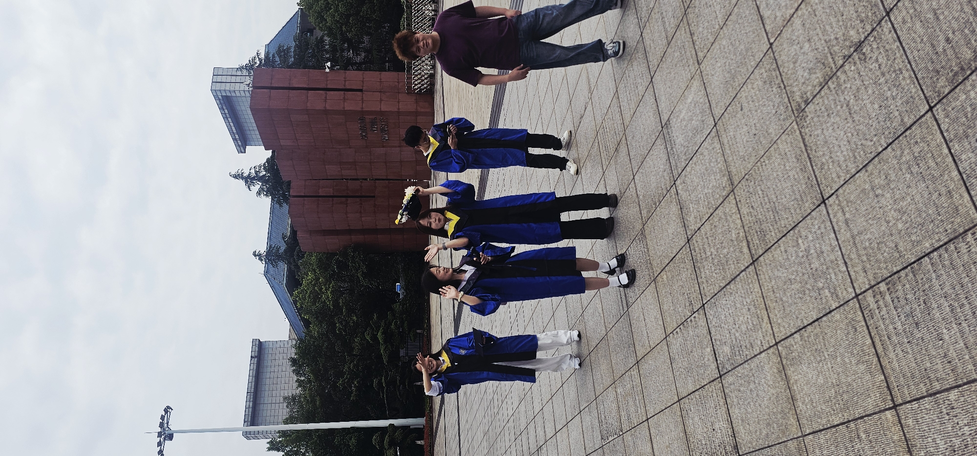 The image, correctly oriented, depicts a joyful outdoor scene featuring five individuals, likely graduates, on a university campus. Four of the people are wearing blue and yellow graduation gowns, while one man is dressed casually.
In the foreground, three female graduates are posing. The woman on the far left, wearing white pants under her gown, holds her hands playfully near her face. Beside her, another woman in a gown and black shoes gestures with open hands. The third woman, positioned slightly behind the other two, holds a graduation cap adorned with flowers and smiles at the camera. A male graduate in a gown stands further back, looking down. On the far right, a man in a purple t-shirt, blue jeans, and sneakers looks towards the camera with a subtle smile.
The setting is a spacious outdoor plaza paved with light-colored, textured tiles. In the background stands a large, reddish-brown monument or building, clearly displaying the vertically oriented text: "CHONGQING NORMAL UNIVERSITY". Lush green trees flank the monument, and modern buildings with glass facades are visible in the distance. The sky is bright and overcast, indicating daytime with diffused light. A tall white pole with light fixtures is visible on the far left. The scene captures a celebratory moment, presumably a graduation ceremony or photo session, at Chongqing Normal University in the Chongqing High-tech Zone, China.