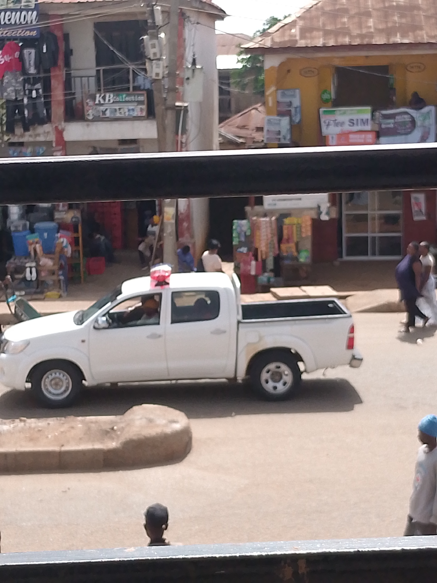 The image depicts a street scene in Bukuru, Nigeria, likely during daylight hours. A white pickup truck with a red and blue emergency light bar on its roof is parked on the right side of the road. A person is visible in the driver's seat. The truck appears to be a commercial vehicle, possibly for security or delivery.
In the background, there are several buildings that seem to house shops. One building on the left has "KB Collection" written on a sign and displays clothing items. Another building on the right has a sign that reads "Free SIM" and is stacked with boxes. People are walking on the sidewalk and street, suggesting a typical day in a busy commercial area. The architecture and the general atmosphere suggest a warm, possibly humid climate. The overall impression is of a lively, functional street environment.