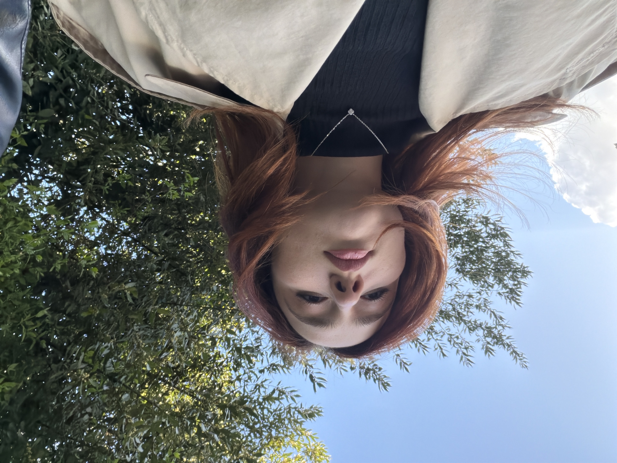 A female individual is depicted in a diagonally oriented outdoor portrait, believed to be taken within the city of Strasbourg, France. Her head is positioned towards the left of the frame, with her body extending towards the lower right. She has long, reddish-brown hair, parted on the side, framing her face. She is wearing a black garment under a light beige trench coat, the collar of which is visible. A thin, silver-colored chain necklace with a small, circular pendant rests on her chest. Her eyes are open and directed upwards towards the camera, and her lips appear lightly colored. The background behind her upper body and head consists of dense green tree foliage, indicating an overhead canopy. The area behind her lower body and to the left displays a clear blue sky with scattered white cumulus clouds. The scene is brightly illuminated by natural sunlight, suggesting daytime.