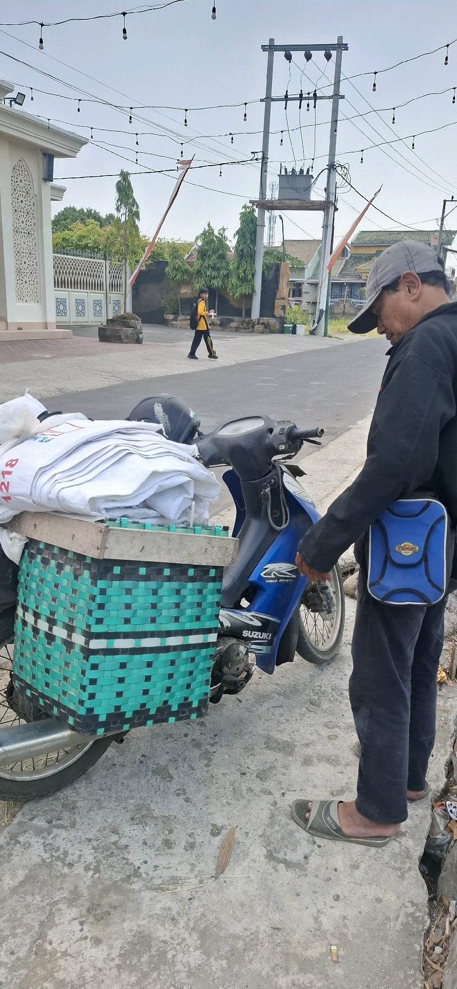 In Paulan, Indonesia, a man wearing a grey baseball cap, black jacket, and dark pants stands next to a blue motorcycle. The motorcycle is equipped with a woven green and black basket on the rear, filled with white sacks. The man is reaching towards the motorcycle. In the background, a person in a yellow shirt and black pants with a yellow backpack walks away from the camera. To the left, a white building with decorative arches and a gate is visible. Overhead, a power line with lightbulbs strung along it stretches across the frame. The scene appears to be a street or roadside with concrete paving.