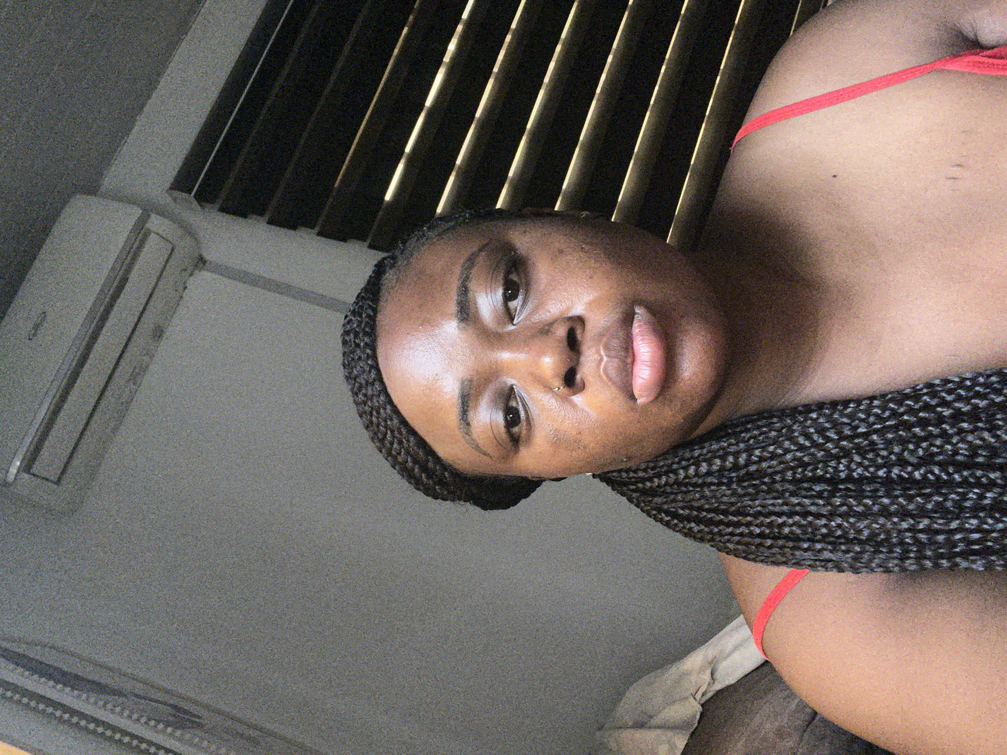 A medium shot shows a Black woman indoors. She has dark hair styled in braids. Her upper body is visible, and she wears a red strap top. The background includes closed blinds and an air conditioner unit. The image lacks detail required to specify the location as Abuja, Nigeria.