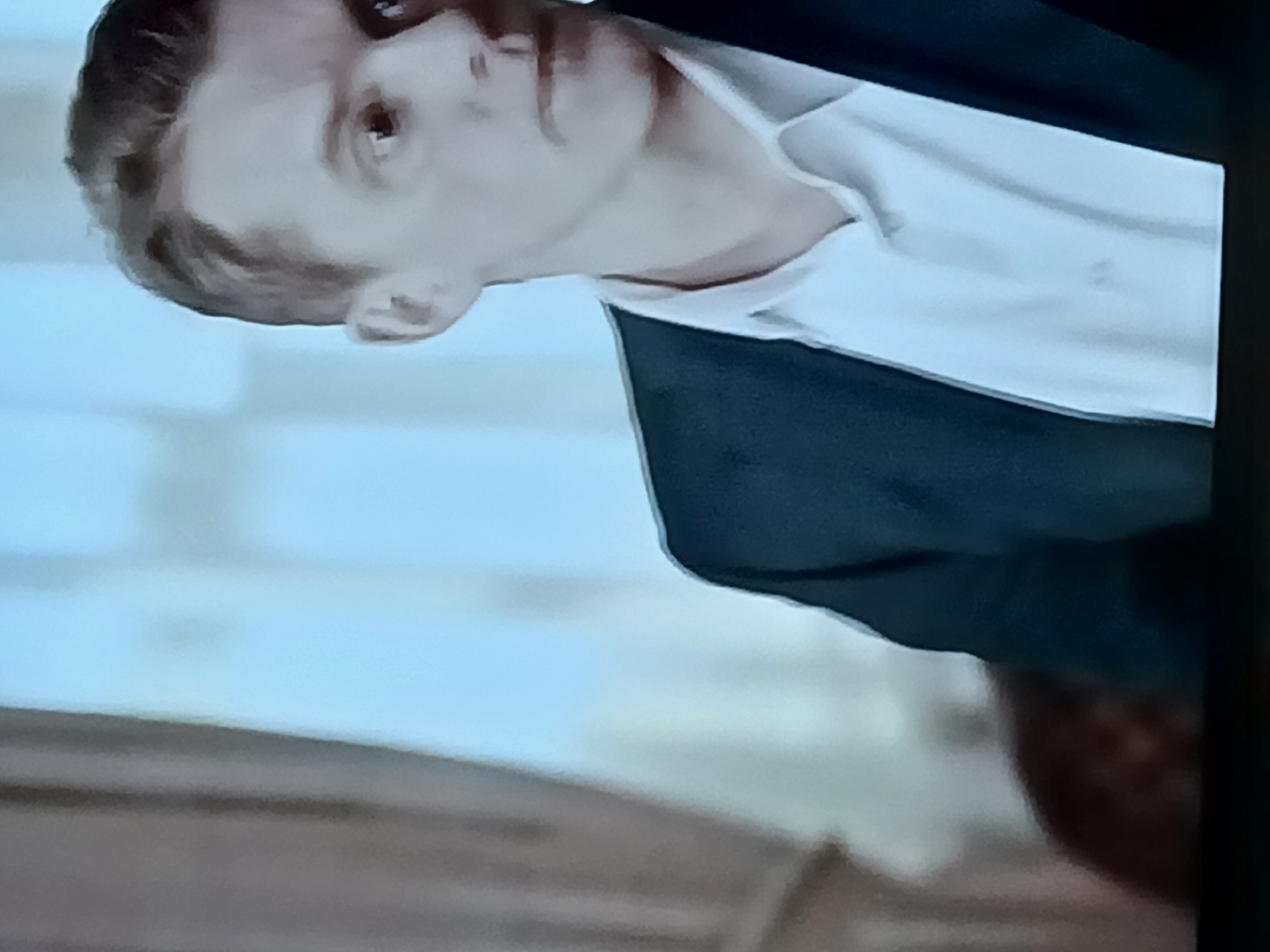 This is a close-up, waist-up shot of a Caucasian male with fair skin and light brown hair. He is wearing a dark suit jacket over a white dress shirt. His hair is styled neatly and appears short. His face is turned to the right of the frame, with his gaze directed outwards. The background is blurred, with a large area of light blue and a darker, textured surface on the left. No specific location in the city of Tara, Russia, can be identified from the image.