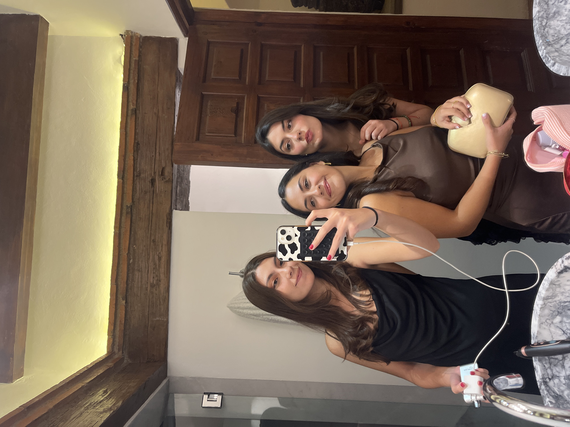 Three young women are taking a selfie in a bathroom with exposed wooden beams and a rustic wooden door. The woman in the foreground on the left is wearing a black dress and holding a portable charger. The woman in the middle is wearing a brown dress and holding a light-colored clutch bag. The woman on the right is wearing a brown top and looking away from the camera. The bathroom appears to be located in a building with traditional Mexican architecture. The presence of the charger and makeup bag suggests the women are preparing to go out or have just returned from an event. The lighting in the bathroom is warm and ambient, likely from recessed ceiling lights.