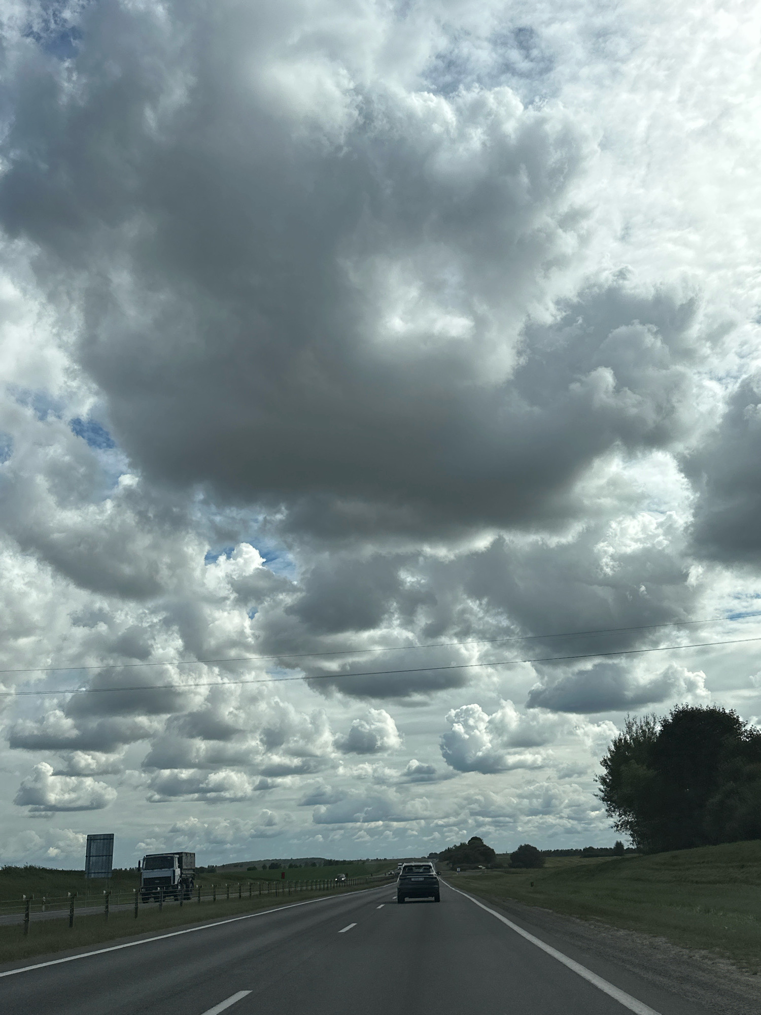 The image captures a highway scene under a dramatic, cloudy sky. In the foreground, a dark-colored car is seen driving away from the camera on the right lane of a paved road. To the left, another vehicle, a light-colored truck, is also moving on the highway. A large, empty sign is visible on the left side of the road, behind a fence. The landscape is characterized by green, rolling hills and scattered trees. The sky is filled with a vast expanse of dense, gray clouds, with occasional patches of lighter clouds and hints of blue sky peeking through. The scene suggests travel through a rural area of Блонскі сельскі Савет, Belarus, with an overcast weather condition.