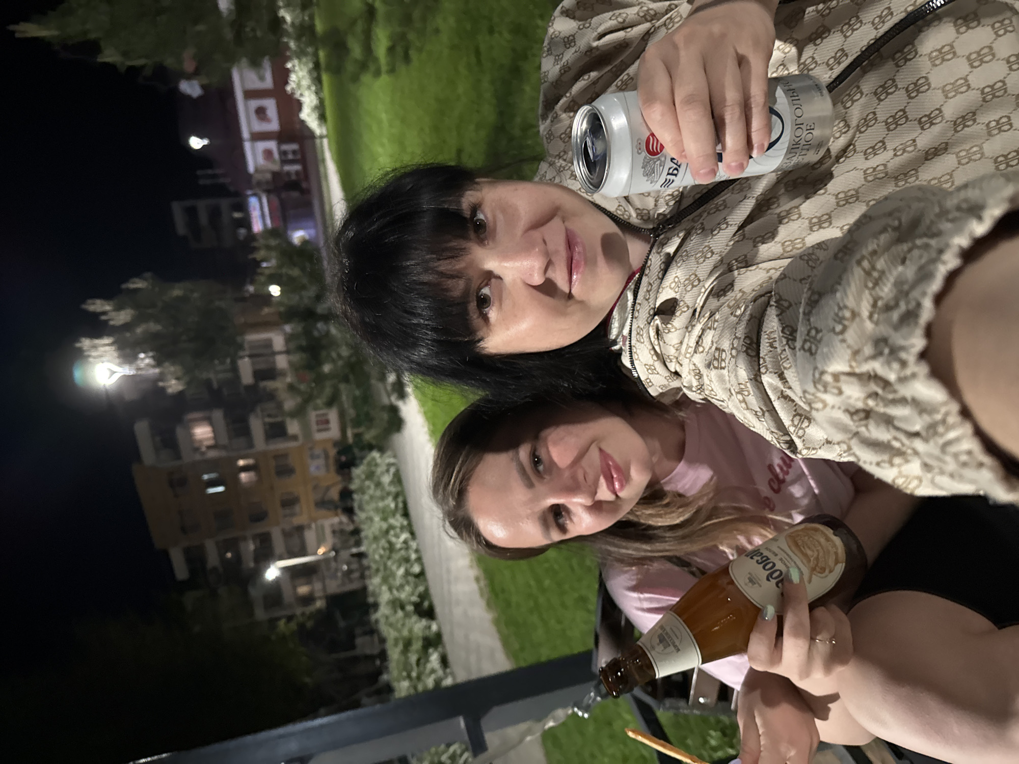 Two women are posing outdoors at night, possibly in Zav'yalovo, Russia. The woman on top, with dark hair, is holding a white can of beer with Cyrillic script. The woman on the bottom, with light brown hair, is holding a bottle of "Dogovar" beer, and pouring beer on the street. Behind them is a residential area with lights from buildings, and some trees.