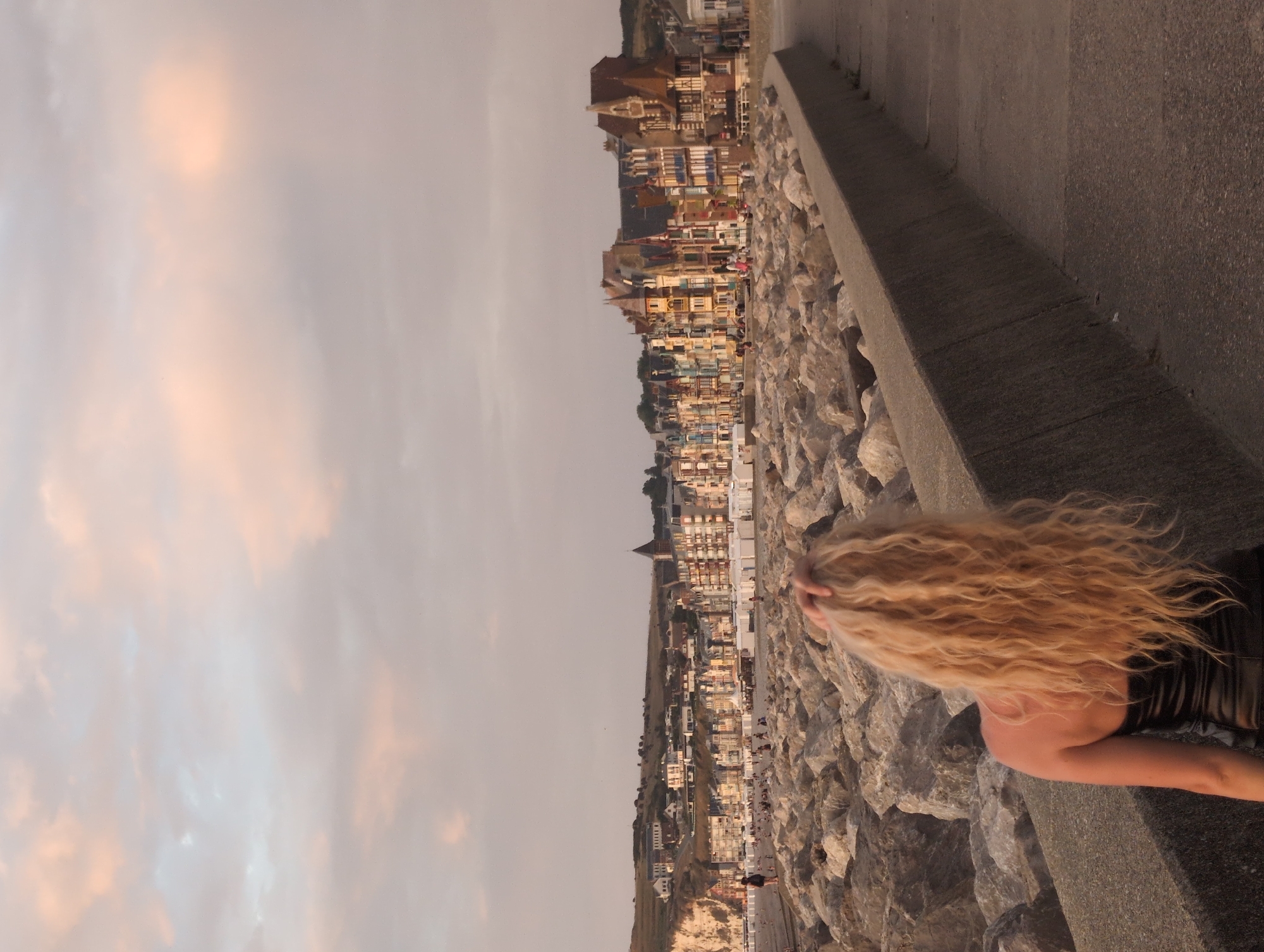 A woman with long, blonde, wavy hair, facing away from the camera, stands on a concrete walkway. She is wearing a black strapless dress. Her right hand is raised, touching her hair. The walkway is lined with a rough, grey concrete barrier, behind which is a large pile of light-colored rocks. In the background, a town with a densely packed row of multi-colored buildings lines the coast. The buildings have varying rooflines and architectural styles, with some featuring ornate detailing. Beyond the town, a green, hilly landscape rises. The sky above is a pale pink and grey, with soft clouds. The scene appears to be a coastal town at sunset. The buildings in the background are characteristic of the Belle Époque architecture found in seaside resorts like Le Tréport, France.