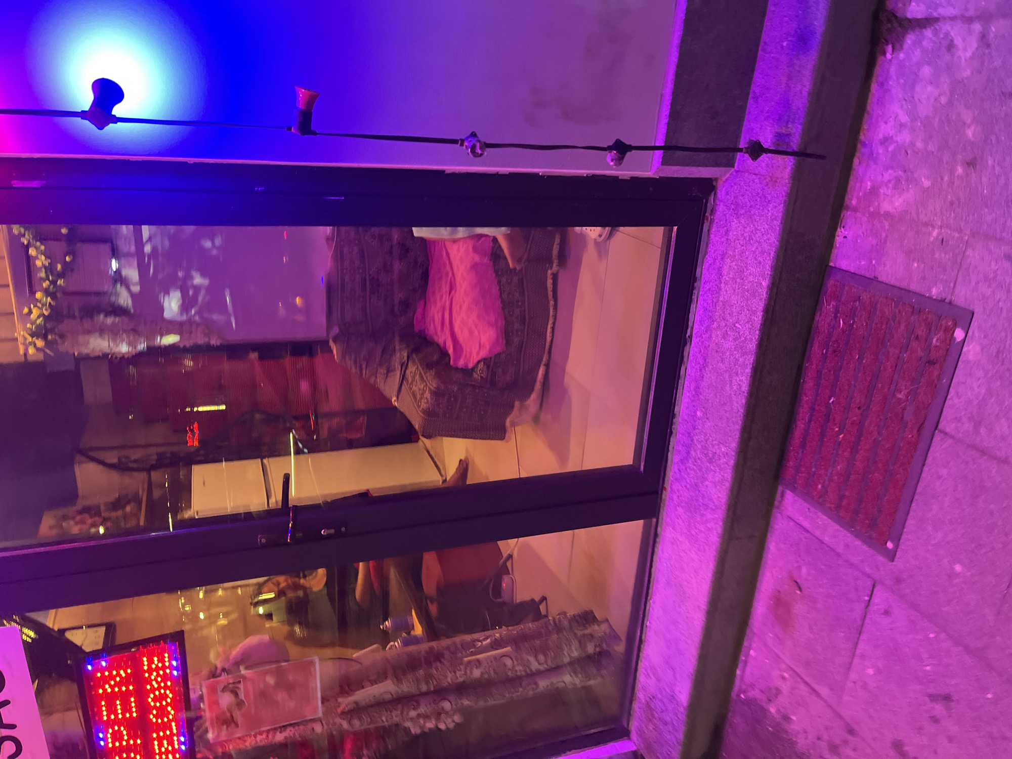 This image captures a view of a storefront at night, bathed in purple and blue neon lights. Through the glass door, a massage parlor is visible, with a woman in casual clothing sitting on an armchair. The interior appears to be decorated with patterned fabrics and a pink cushion on the chair. A sign above the door reads "OPEN WELCOME" in red LED lights. Outside, a red doormat lies on the pavement. The establishment is likely located in Batumi, Georgia, a popular tourist destination known for its nightlife and entertainment venues.