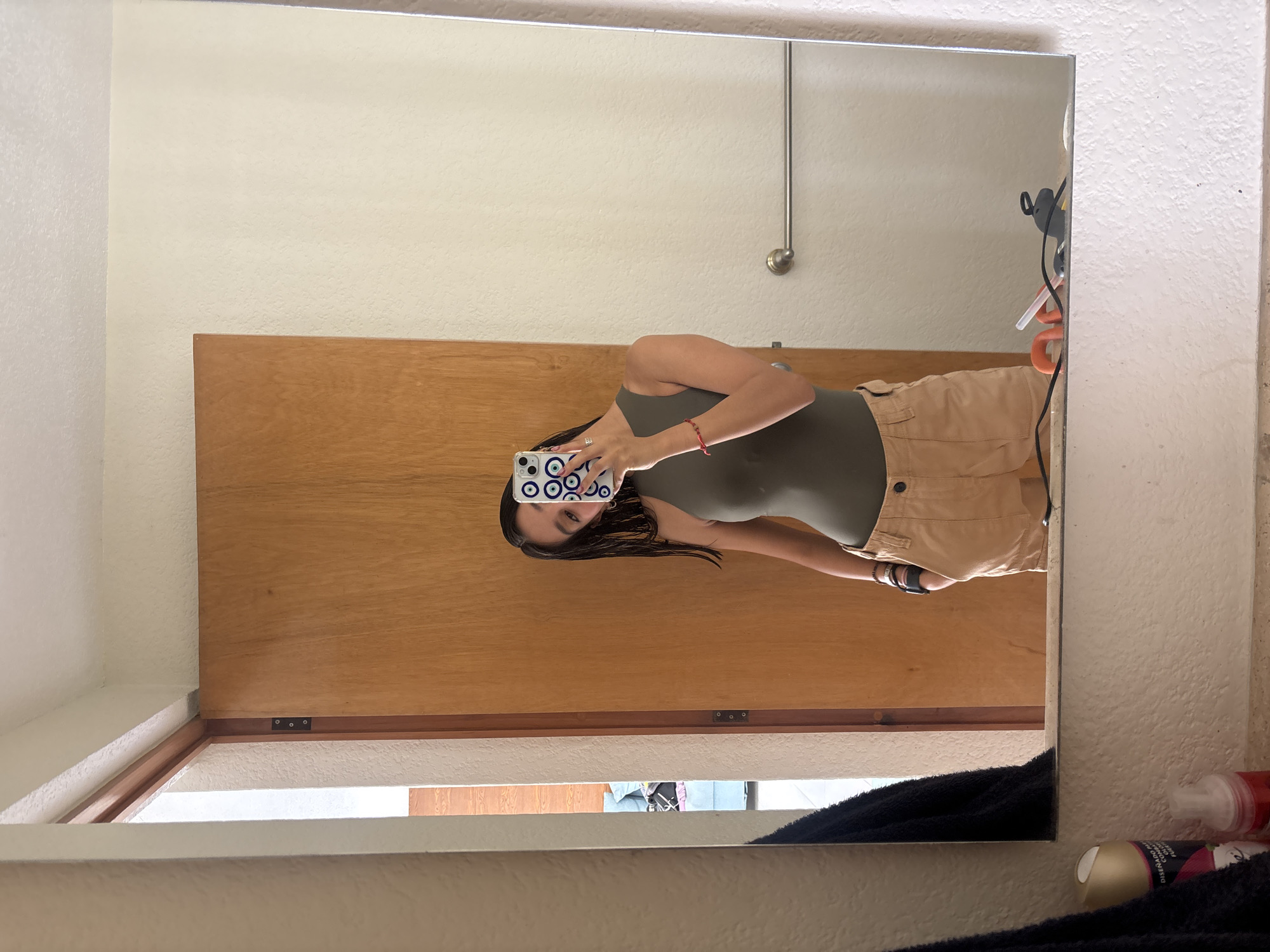A person stands in front of a mirror, reflected within. The mirror is surrounded by a white frame, and the reflected background includes a light beige wall and a brown wooden door. A metal pipe fixture is mounted on the wall. A black towel and various toiletries clutter the bottom edge of the mirror. The person is dressed in a green top and khaki shorts, holding a smartphone with a blue and white case up to take the photo. No information indicates the location is in the city of Puebla, Mexico.