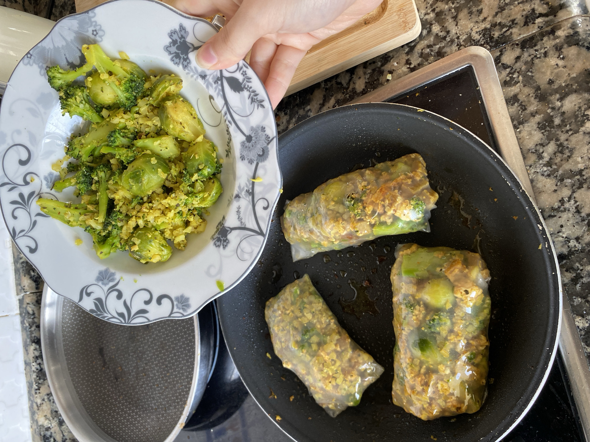 A close-up, eye-level shot captures a cooking scene in a kitchen. The main subjects are a plate of cooked vegetables, a hand holding the plate, and a frying pan containing wrapped food items.
In the left foreground, a white plate with an intricate gray floral and swirl pattern holds a generous serving of bright green, cooked vegetables. These include visible florets of broccoli and halved brussels sprouts, mixed with a finely crumbled, yellow-hued substance, possibly seasoned cauliflower rice or another grain.
A fair-skinned hand, with a silver ring on one finger, gently holds the edge of this plate, positioning it over a black non-stick frying pan situated on a stovetop. Inside the pan, three rectangular rolls are cooking. These rolls are wrapped in a translucent, thin material, likely rice paper, allowing the green vegetable pieces (identifiable as broccoli florets) and a textured, yellowish-brown filling to be seen clearly within. A small amount of oil or cooking liquid is visible at the bottom of the pan.
The background reveals a speckled dark granite countertop, a section of a black stovetop, and a wooden cutting board partially visible in the upper right corner. The lighting suggests an indoor, likely daytime, setting. The scene depicts an activity of food preparation, possibly assembling or cooking spring rolls filled with a vegetable and spiced mixture, with a side of similar cooked vegetables. No discernible text is visible in the image.