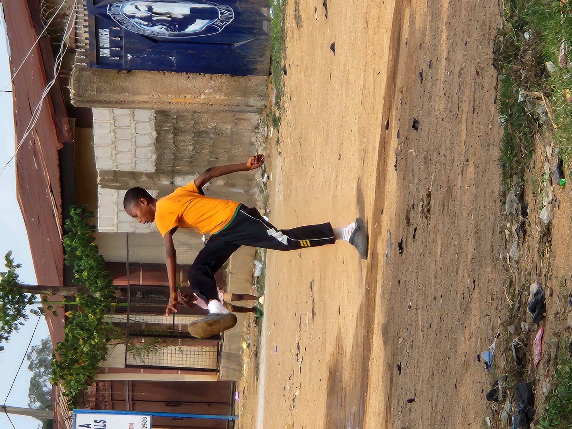 A young boy, wearing an orange t-shirt and black athletic pants with yellow stripes, is captured mid-action, appearing to kick or punt a large, round object resembling a rock or a bundled piece of cloth. He is in a dusty, outdoor setting, possibly a street or field. His body is contorted, with his right leg raised and bent, and his left leg planted firmly on the ground. His arms are outstretched, one pointing forward and the other slightly bent.
The background reveals a building with a red, corrugated metal roof and a blue, ornate gate with a circular emblem. To the left, there is lush green foliage. The ground is primarily a dry, sandy dirt path with scattered debris, including plastic bags and other refuse, particularly along the edge where grass grows.
The lighting suggests it is daytime, with shadows cast on the ground, indicating the sun is out. The overall scene feels informal and possibly rural or suburban. The boy's focused expression and dynamic pose suggest he is engaged in a game or sport. There is a glimpse of what appears to be a sign with text on a blue background at the bottom of the frame, but the text is mostly illegible. The location context of Sabon Tasha, Nigeria, places this scene within a specific geographic and cultural environment.