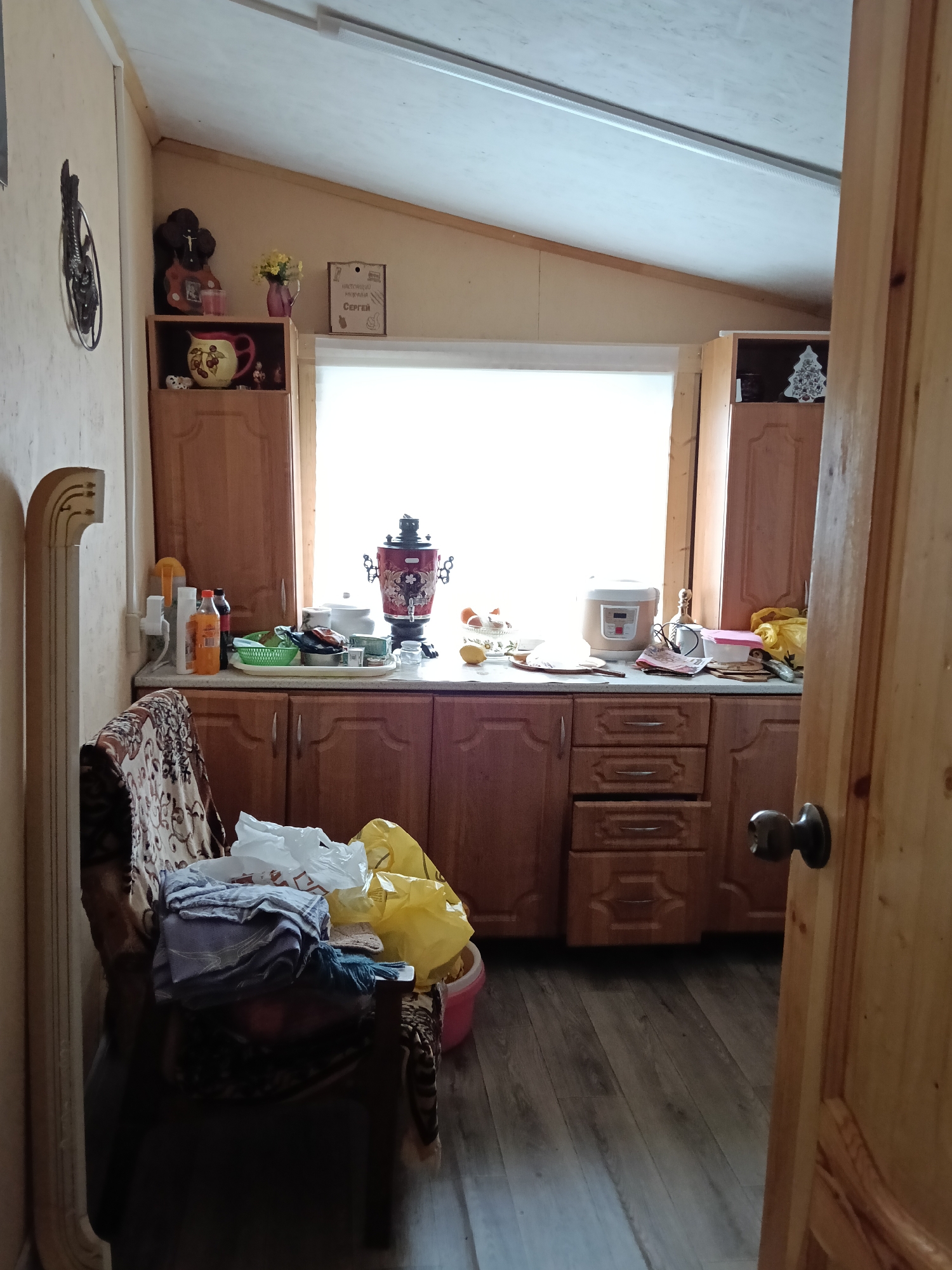 The interior of a cluttered kitchen in a residence in Красный Маяк, Russia. The room features light-colored wooden cabinets and drawers. A countertop holds various items, including a red samovar, a rice cooker, food items, and kitchen utensils. The room is brightly lit by daylight coming in from a window. A chair in the foreground is covered with clothing and plastic bags. A wooden door on the right side of the image is ajar. The floor is made of wood.