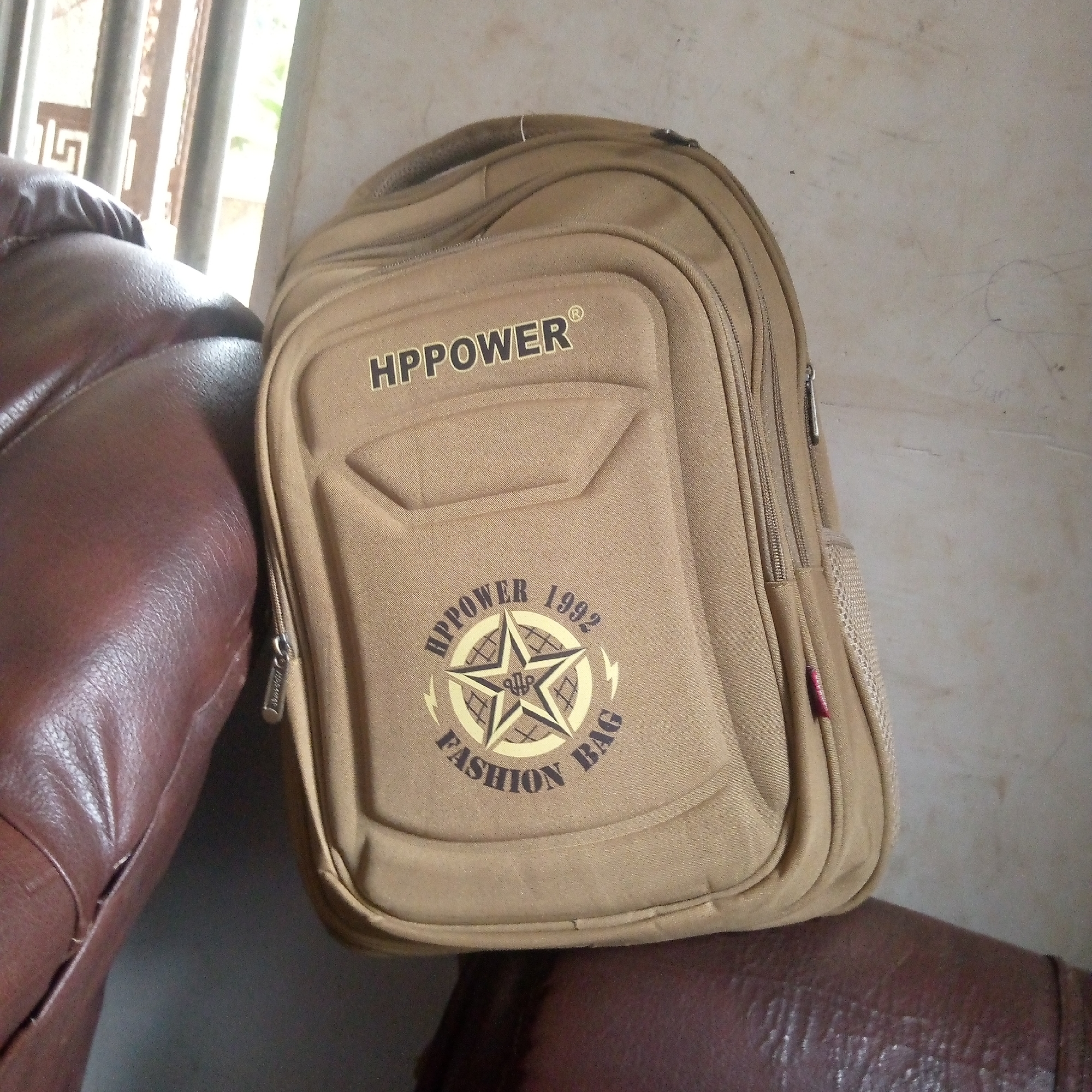 A tan HPPOWER backpack rests on a brown leather surface. The backpack has multiple zippered compartments and a mesh side pocket. The inscription "HPPOWER 1992 FASHION BAG" is displayed on the backpack's front. In the background, a partially visible window with white bars or molding is present, along with a textured beige wall. The location is Unknown, Unknown.